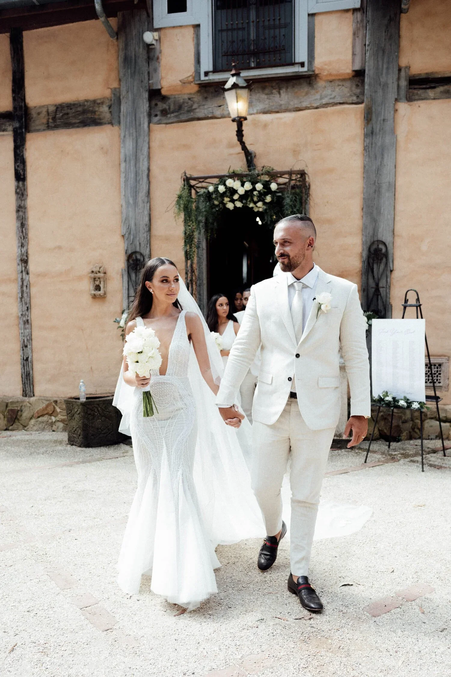 A bride and groom holding hands walking outside during wedding ceremony, with woman in white dress holding bouquet, on rustic building background with flower decoration