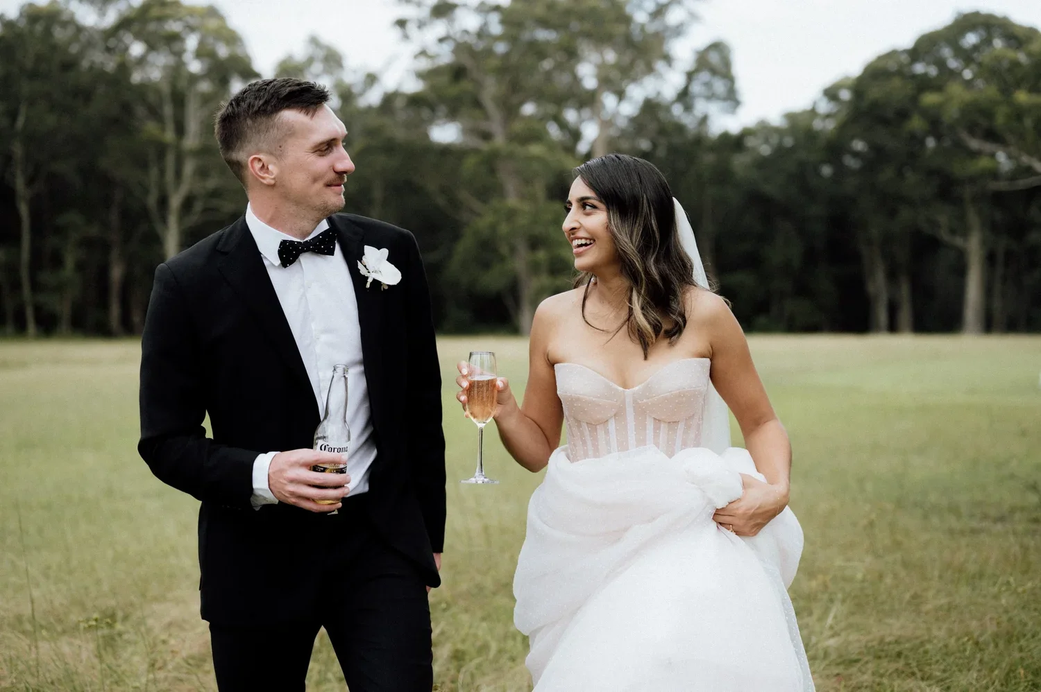 A bride and groom smiling at each other during their outdoor wedding. The groom is in a black tuxedo with a polka dot bowtie, holding a Corona beer bottle. The bride is in a strapless wedding dress, holding a glass of champagne, and carrying the skir