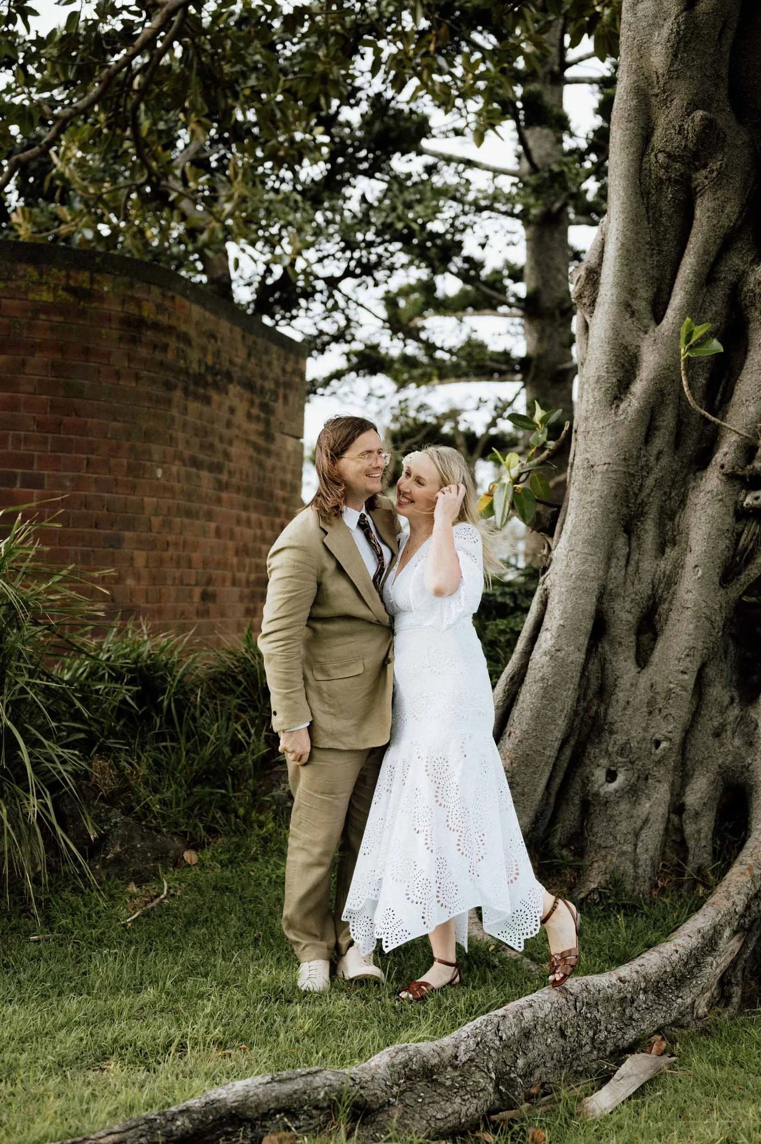 A couple dressed in wedding attire standing outdoors near a large tree, with greenery and a brick wall in the background.