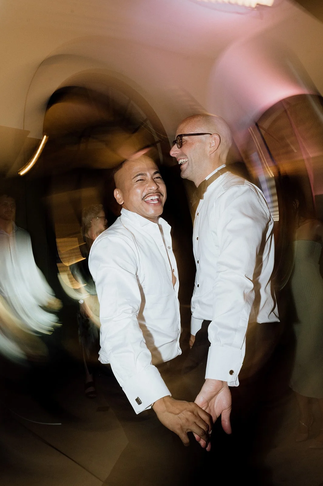 Two grooms on the dancefloor at their same sex wedding in white dress shirts holding hands and laughing at a celebration event.