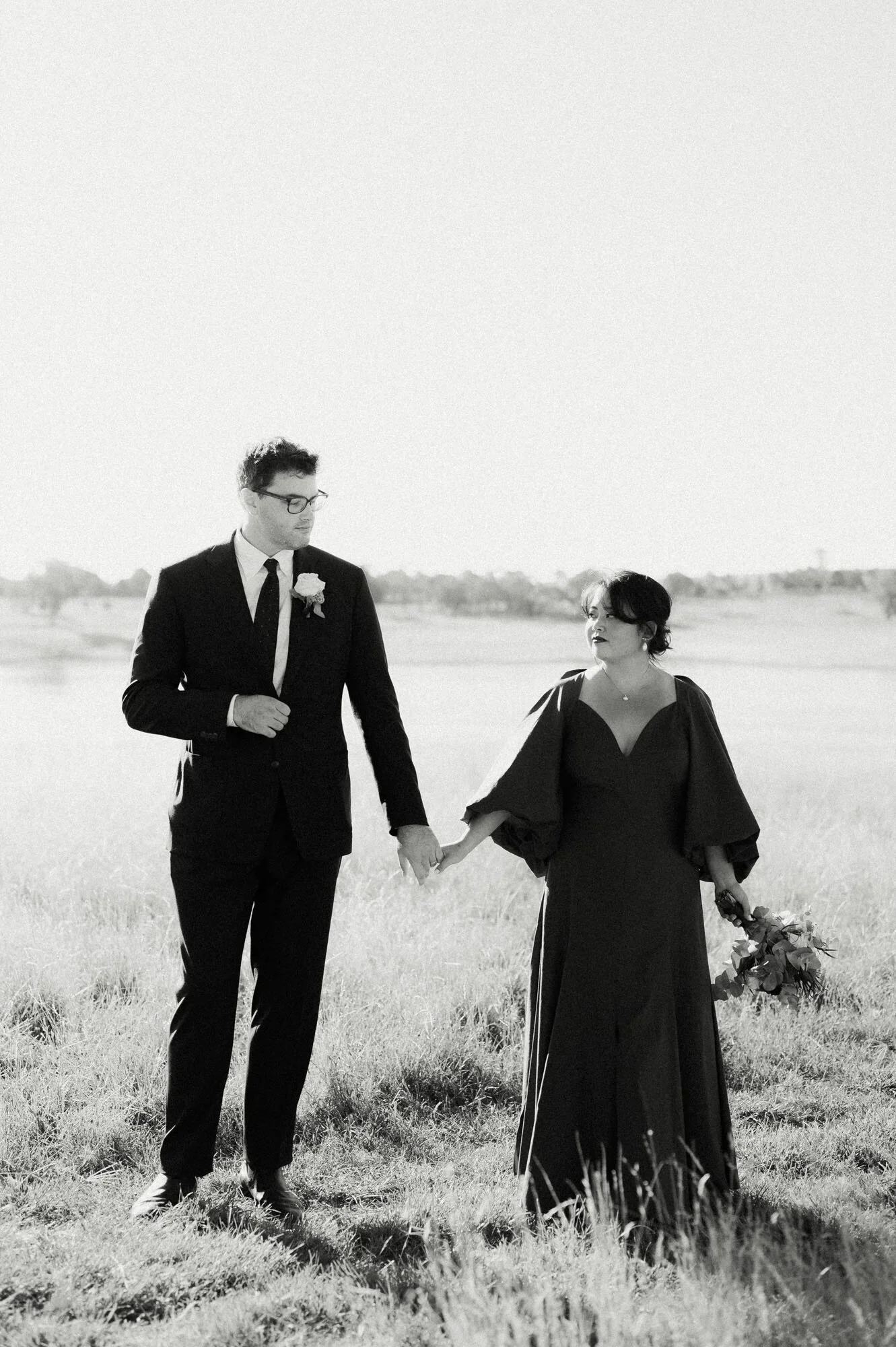 A black and white photo of a couple walking outdoors on a grassy field, holding hands. The man is dressed in a suit with a boutonniere, and the woman is in a flowing dark dress and holding a bouquet.