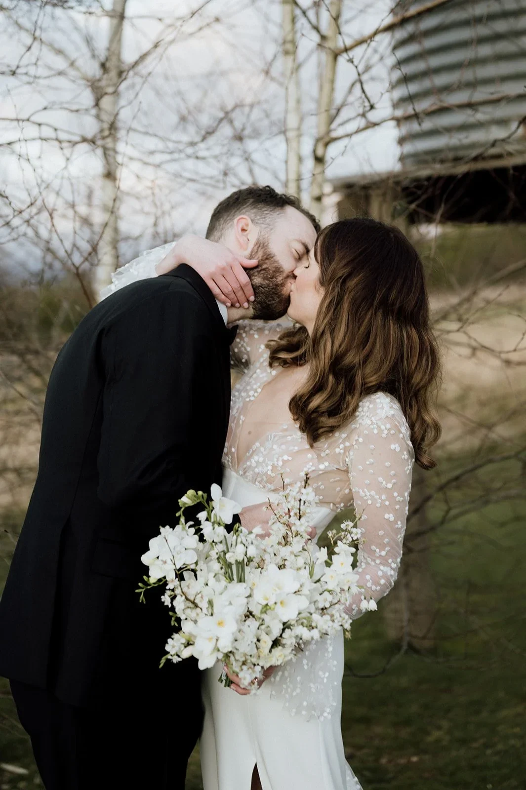 A newlywed couple sharing a kiss outdoors, the woman holding a white flower bouquet, with bare trees and a building in the background.