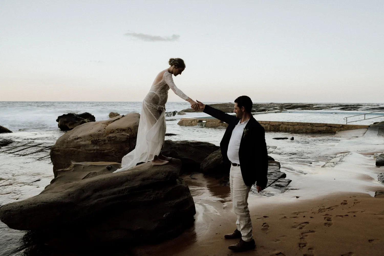 A woman in a white dress standing on rocks at the beach reaching out to a man in a suit who is standing on the sand, holding her hand. The scene is set at sunset or dusk by the ocean.