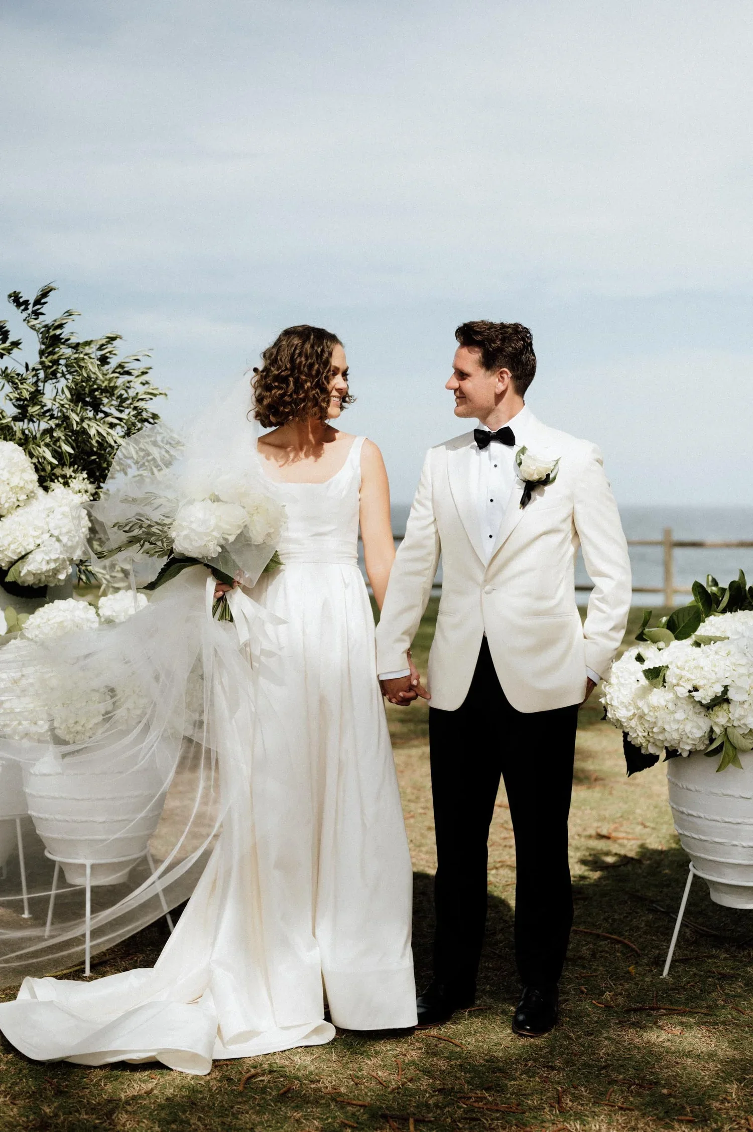 A bride and groom holding hands and smiling at each other outdoors, surrounded by white flowers and greenery, on a cloudy day.