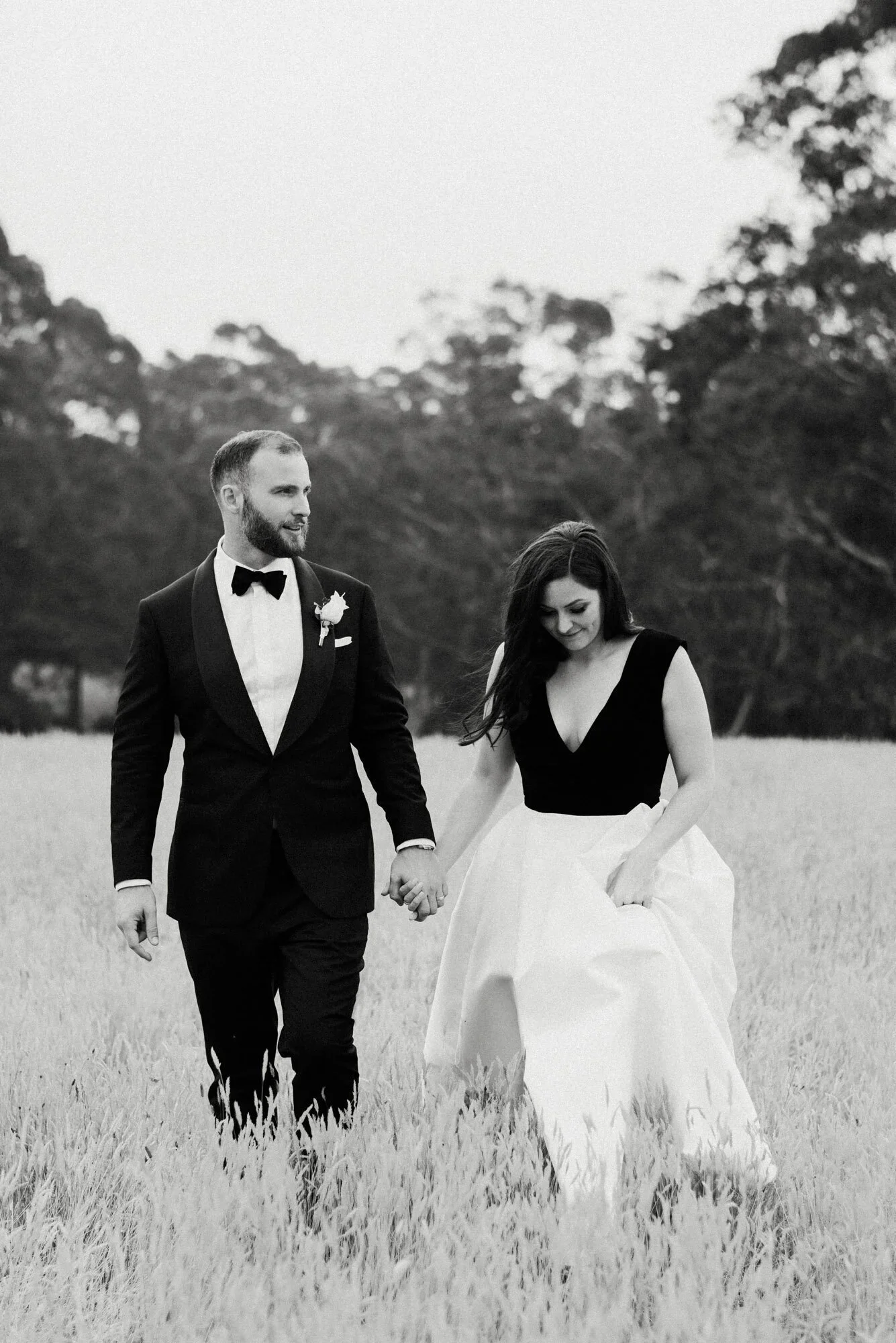 A black-and-white photograph of a couple walking hand in hand through a grassy field, with the woman dressed in a wedding gown and the man in a tuxedo, surrounded by trees in the background - Bowral wedding photographer.