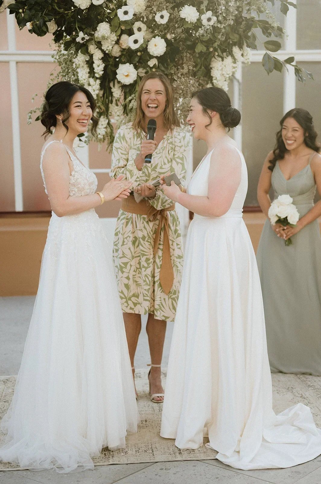 A wedding ceremony with two brides exchanging rings, one with dark hair in an updo and the other with shoulder-length dark hair, standing before an officiant and a bridesmaid, all smiling and laughing, with a floral arch in the background.