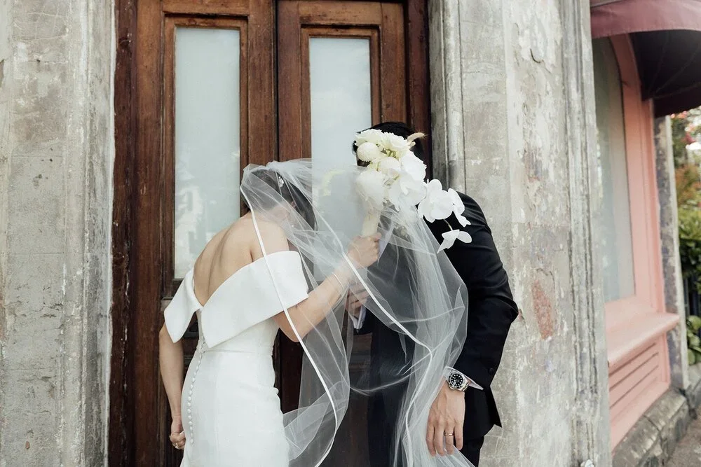 Bride and groom share a kiss behind a bridal veil in front of a wooden door, with the bride in a white off-shoulder dress and the groom in a black suit holding a bouquet of white flowers.