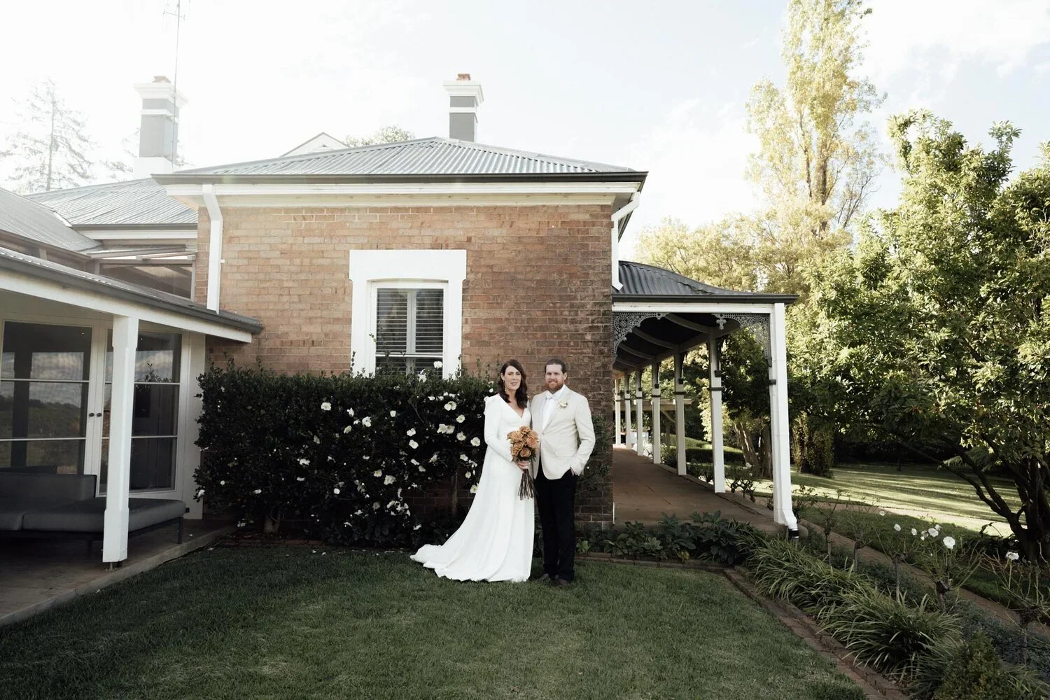 A bride and groom standing outdoors in front of a brick house with white trim, green bushes, and trees, during daytime. The bride is holding a bouquet of flowers and wearing a white wedding dress, the groom is in a white suit jacket. They are smiling