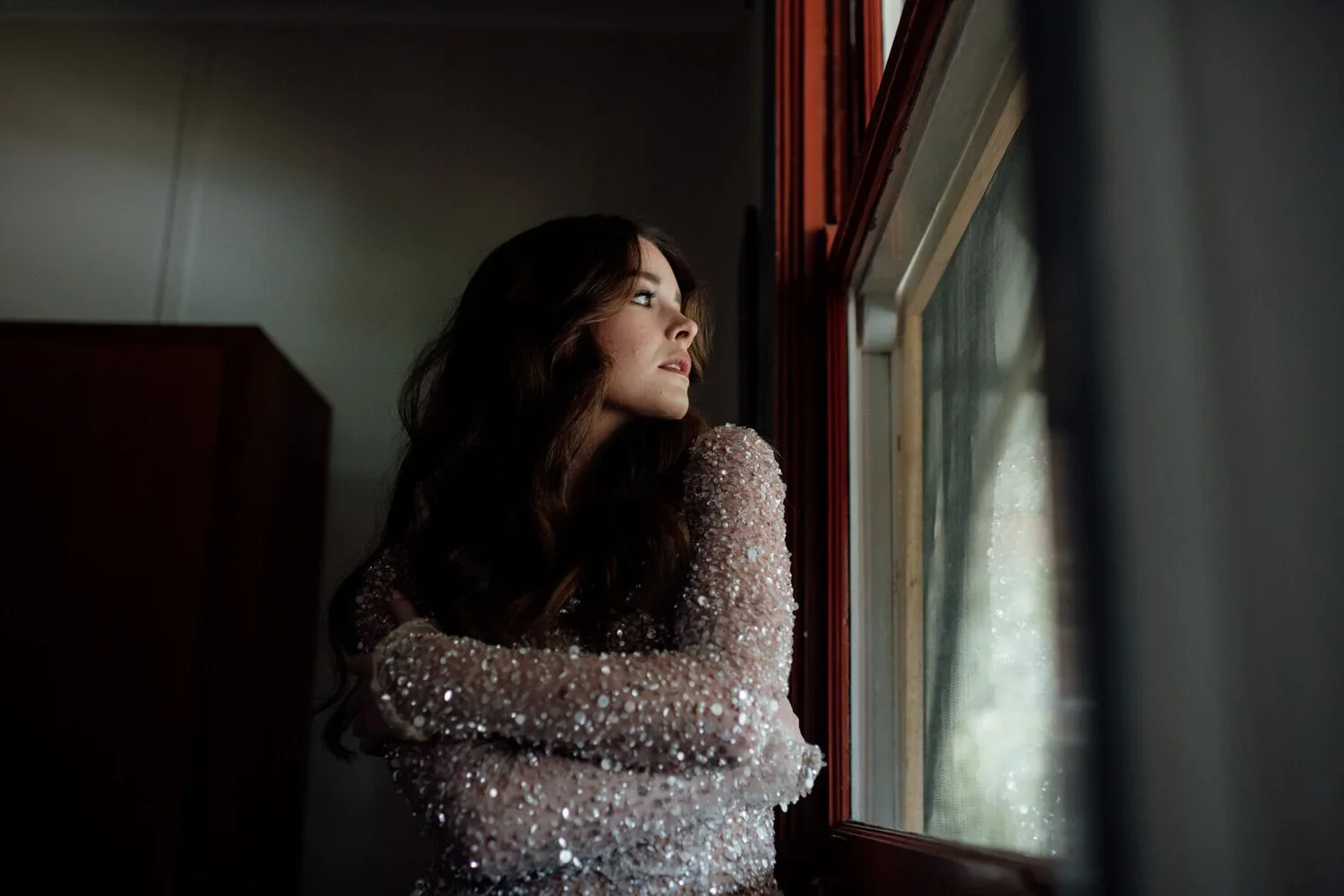 A woman with long dark hair is looking out a window, wearing a sparkling, sequin-covered dress.