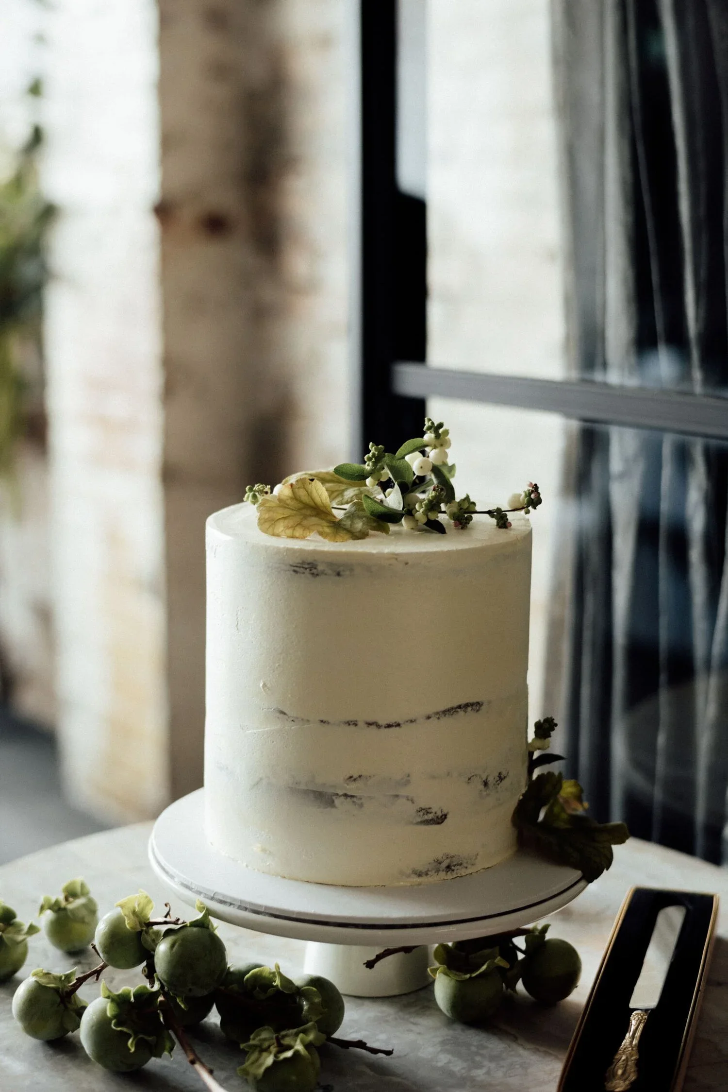 A white, two-tiered vanilla cake decorated with sprigs of green leaves and small white berries, placed on a stand. Fresh green berries and leaves are scattered around the cake on a table with a marble surface. A window with sunlight streaming through