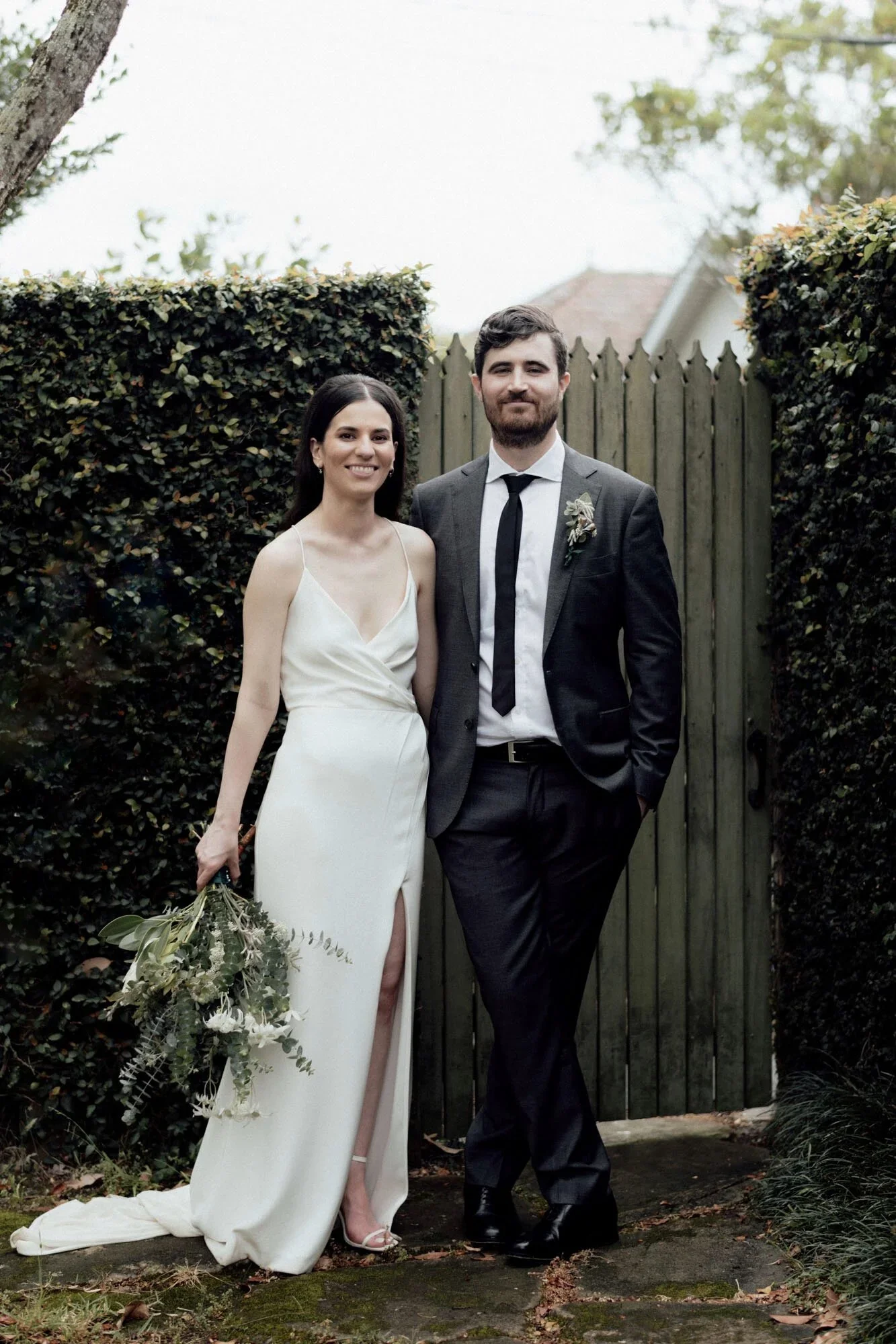 A couple dressed in wedding attire celebrating outdoors in front of a wooden gate and hedge, with a woman in a white gown holding a bouquet and a man in a suit.