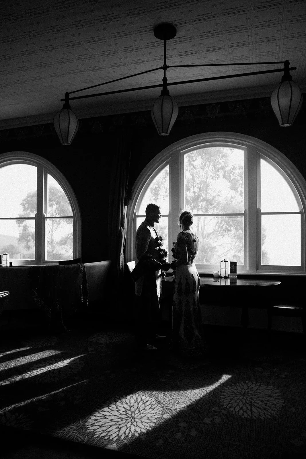 A black and white photo of three people standing by a large arched window, holding hands and engaging in conversation, with outdoor trees visible through the window behind them. The room has a ceiling with decorative panels and a hanging light fixtur