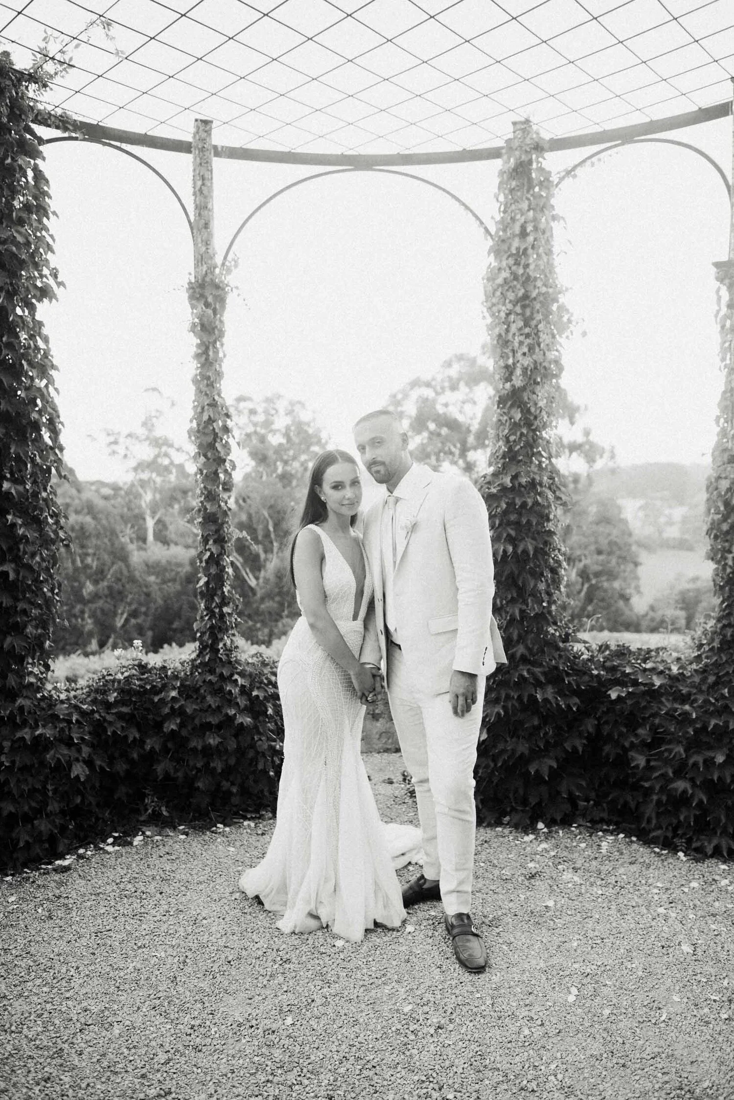 A black and white photograph of a newlywed couple standing outdoors under an arching trellis covered with vines, with trees and an open landscape in the background.