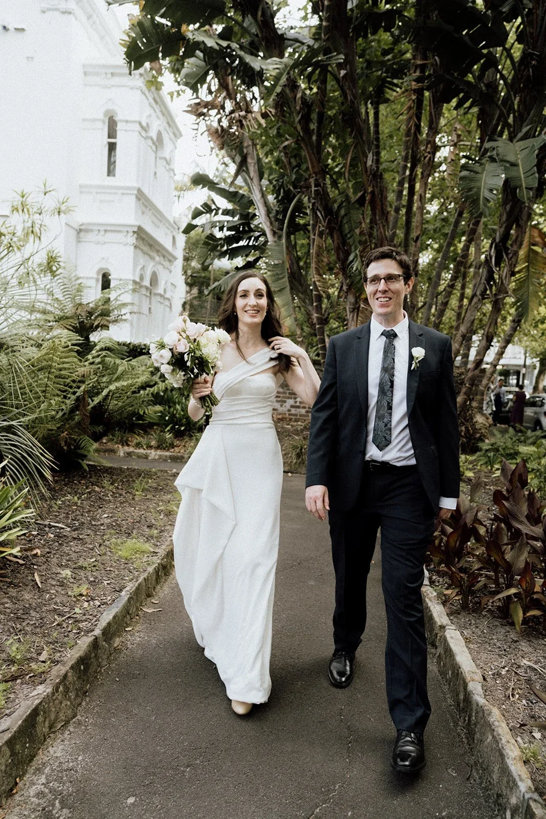 A bride in a white wedding dress holding a bouquet of flowers, walking with a groom in a black suit and tie, outdoors surrounded by greenery, trees, and a white building in the background.
