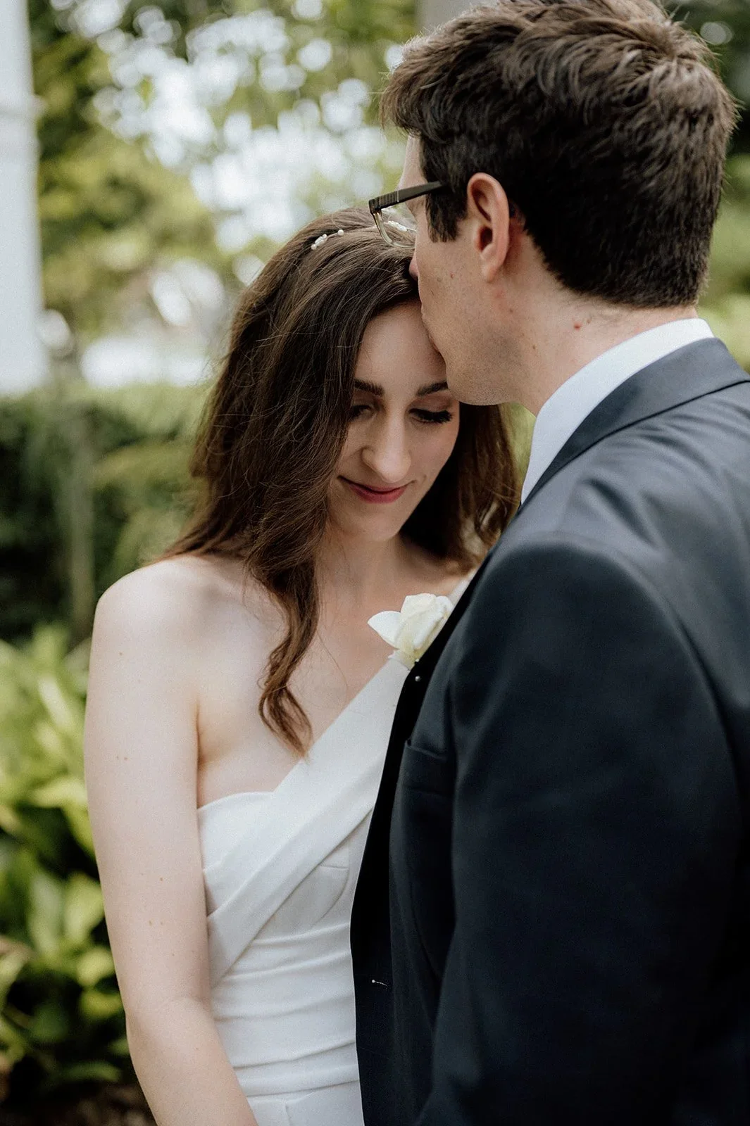 A bride and groom share a tender moment outdoors, with the groom gently kissing the bride's forehead, both dressed in wedding attire surrounded by greenery.
