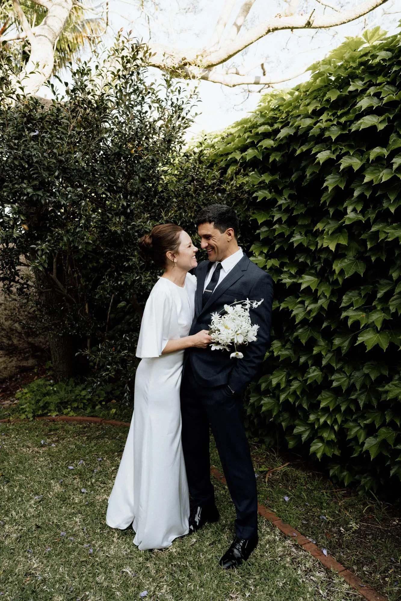 A bride and groom stand close together in a garden surrounded by greenery, smiling and looking into each other's eyes. The bride holds a bouquet of white flowers, and the groom is dressed in a dark suit with a tie. They are sharing an intimate moment