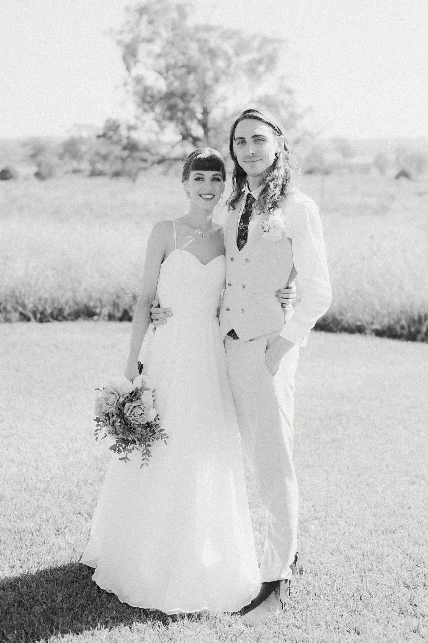 Black and white photo of a bride and groom outdoors on a sunny day. The bride is holding a bouquet of roses, and both are smiling with the groom having his arm around the bride. The background features a tree and open grassy field.