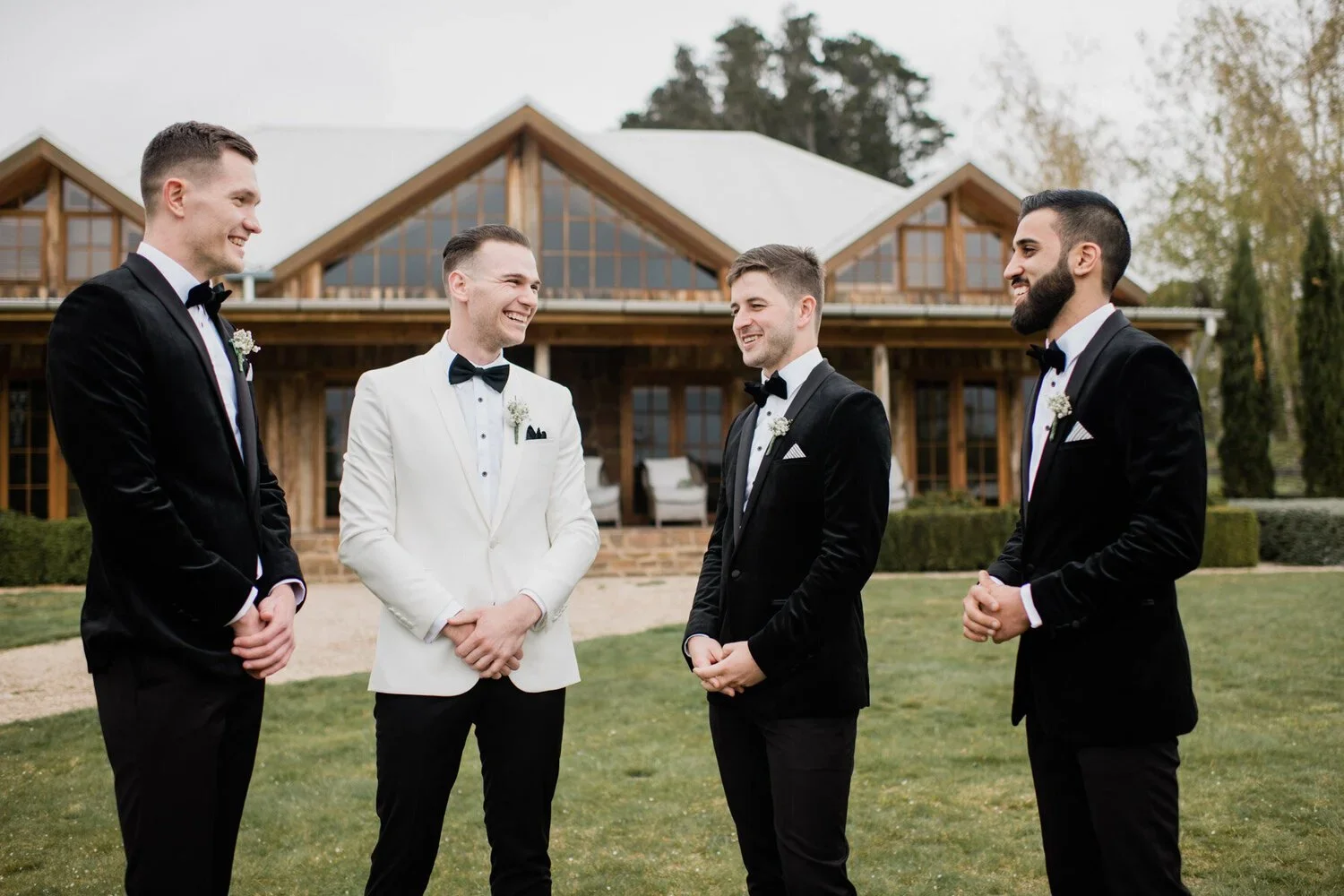 Four men in tuxedos standing outside a house, smiling and talking to each other.