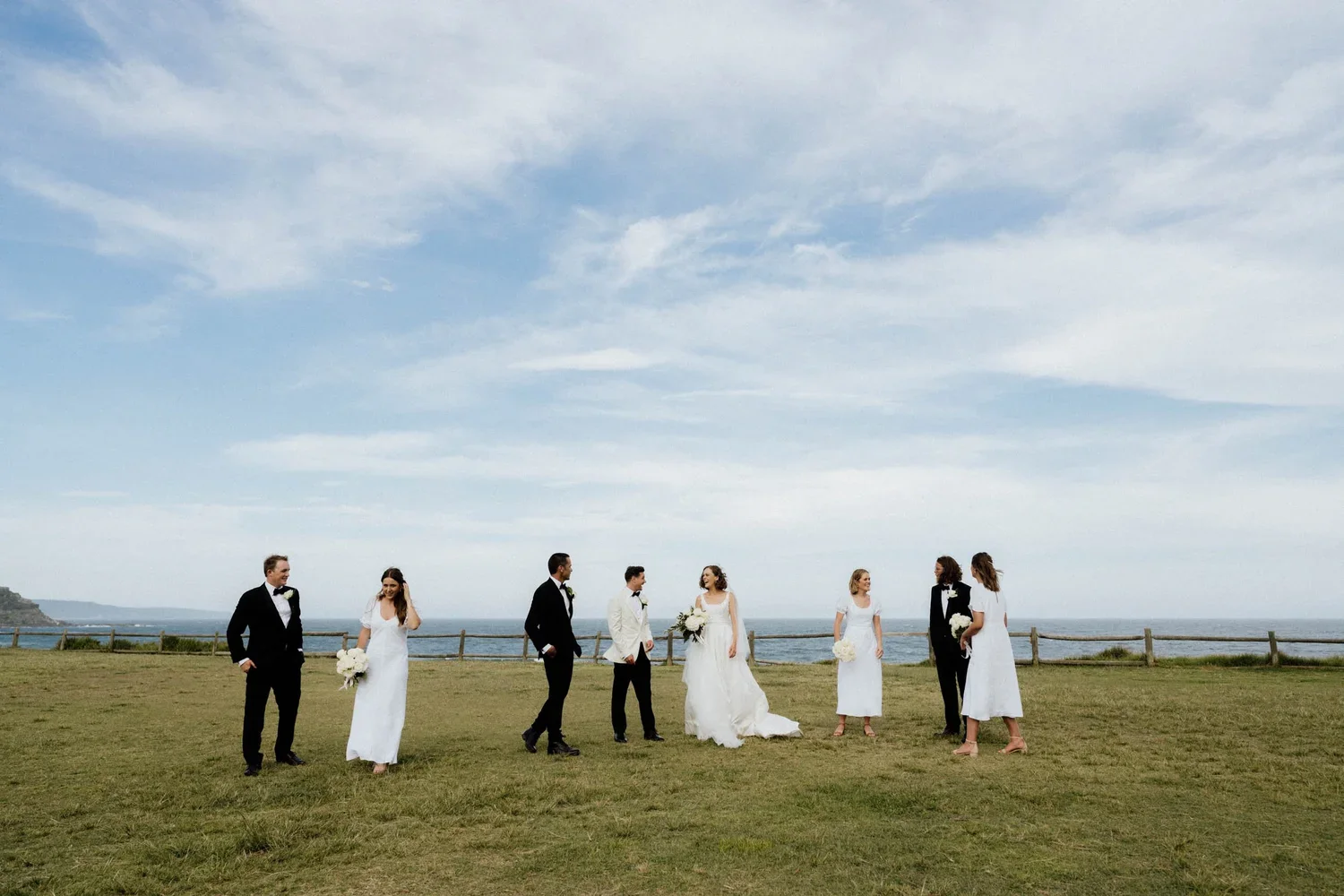 A wedding party on a grassy field near the beach with the ocean and cloudy sky in the background. The group includes brides and grooms, some holding bouquets, dressed in formal wedding attire.