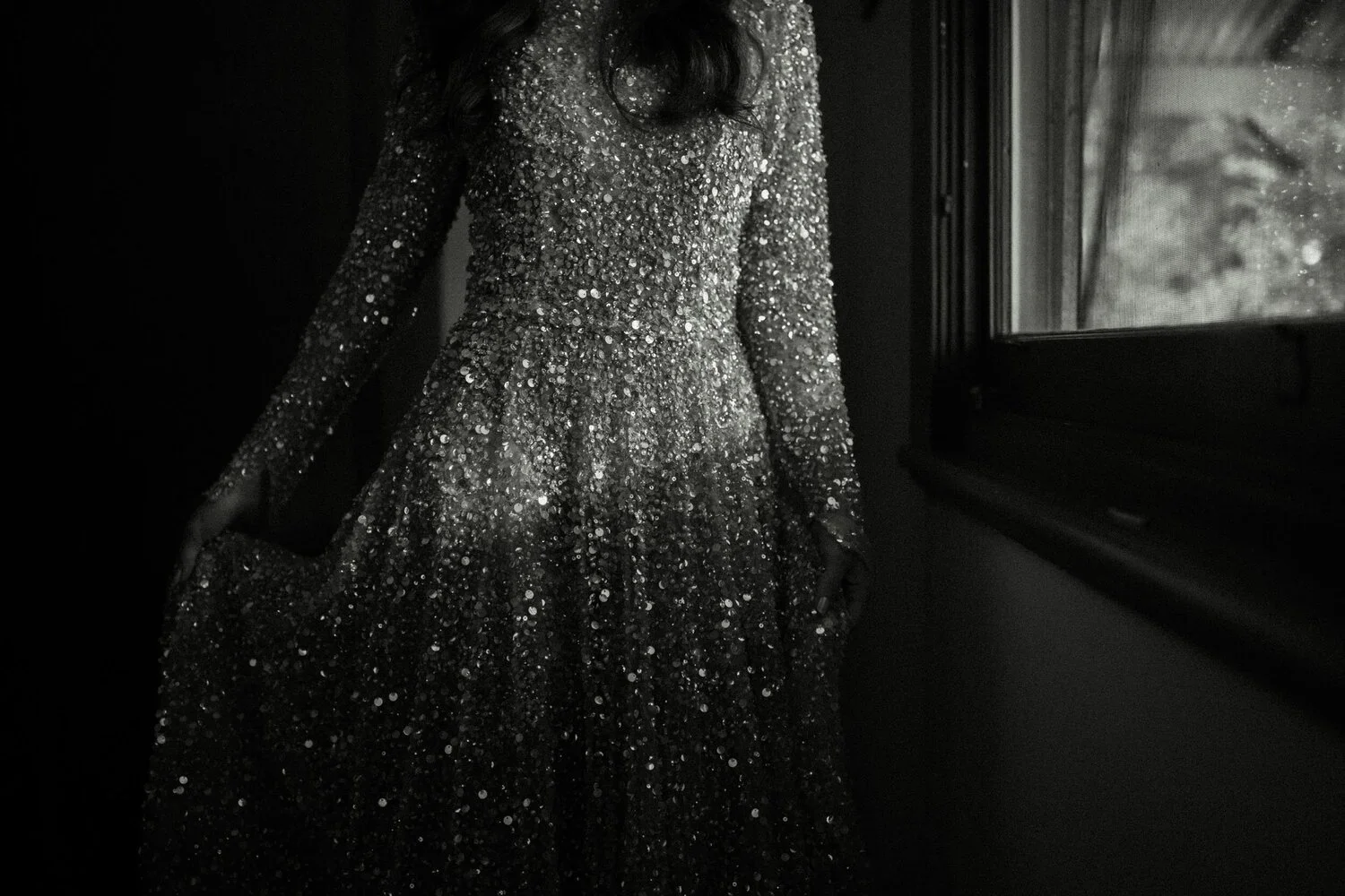 A woman wearing a long, sparkling, sequined gown standing near a window, with natural light reflecting off the dress.