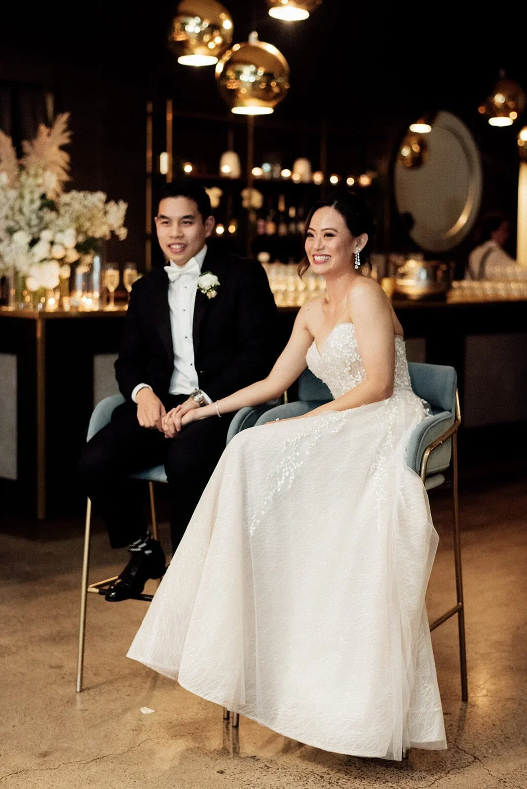 A bride and groom sitting together at their wedding reception, holding hands, smiling, with elegant decor and dim lighting in the background.