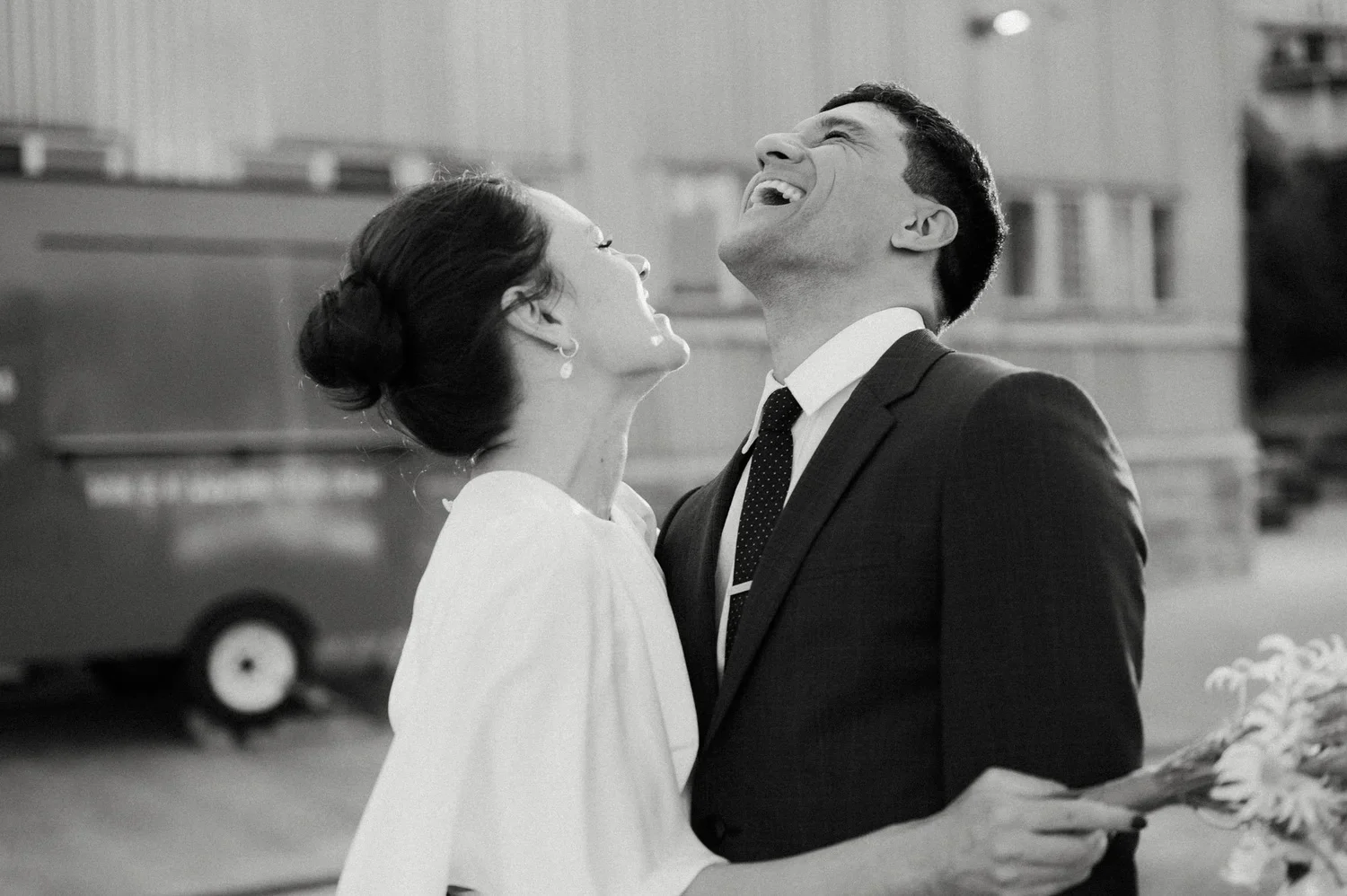 A black and white photo of a joyful couple, a woman in a white dress and a man in a suit, sharing a happy moment. The woman is holding a bouquet of flowers.