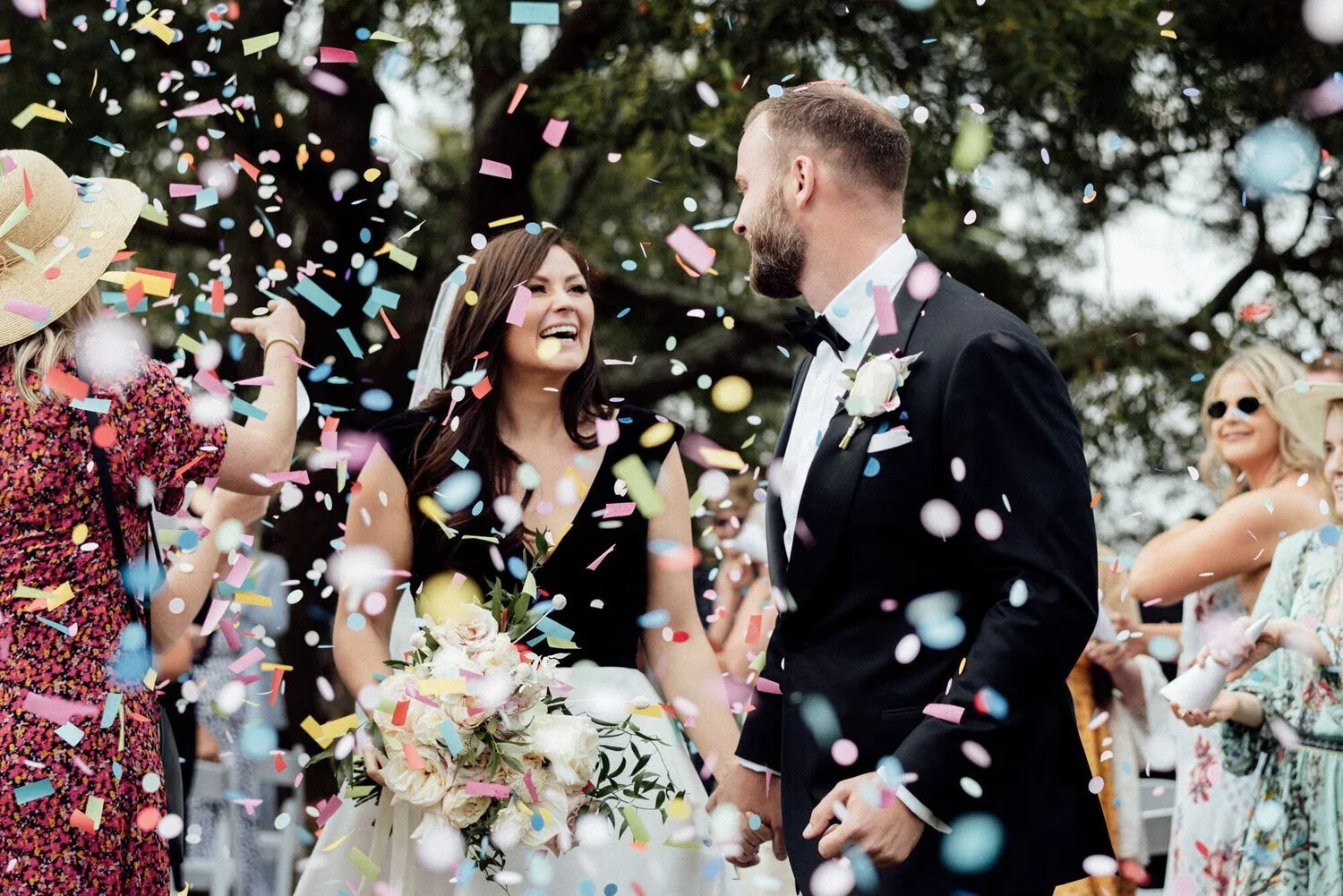 Bride and groom smiling at each other as friends throw confetti during outdoor wedding ceremony.