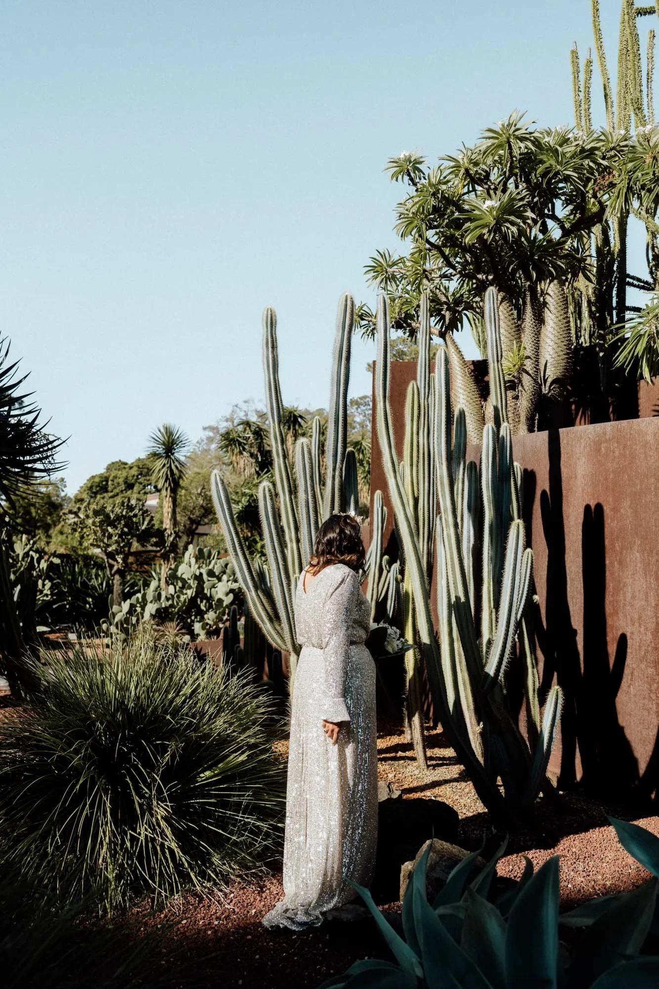 A woman in a sparkly, long silver dress standing among tall cacti and desert plants in a sunny outdoor setting with a clear sky.