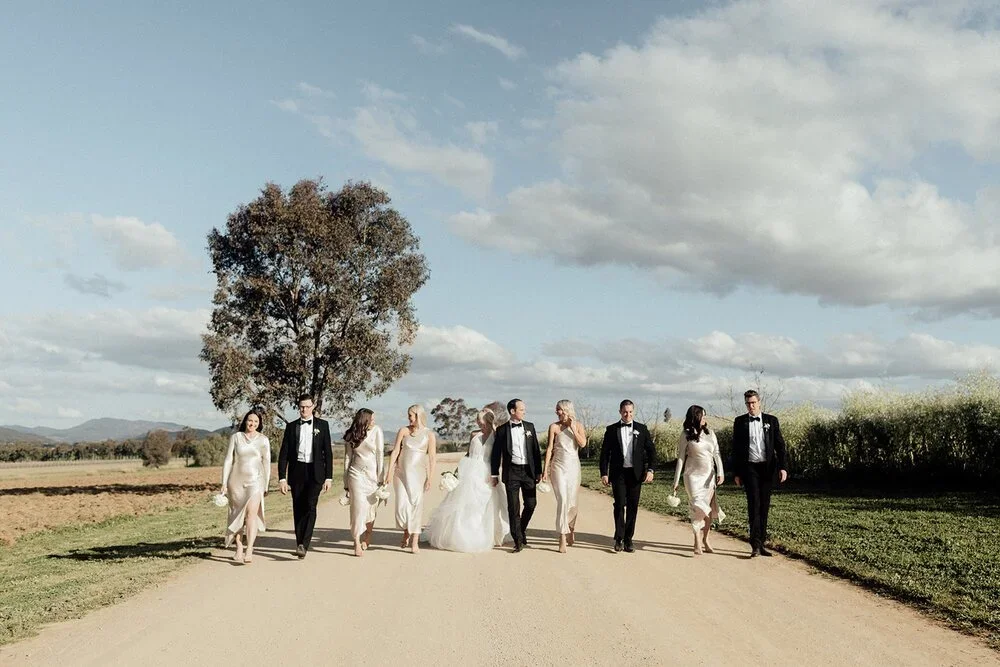 A wedding party walking outdoors on a dirt road with fields and a tree in the background under partly cloudy skies - Mudgee Wedding Photographer.