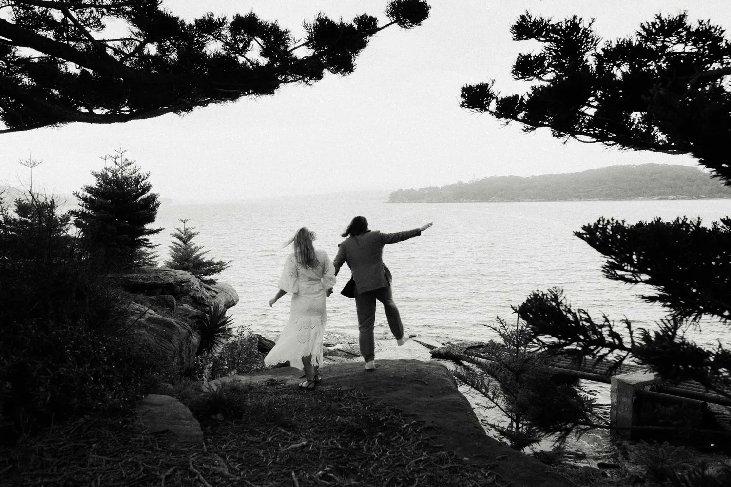 Two people, a man and a woman, walking hand in hand along a rocky shoreline by the water, framed by trees, with the woman wearing a long white dress and the man wearing a suit, gesturing towards the water.