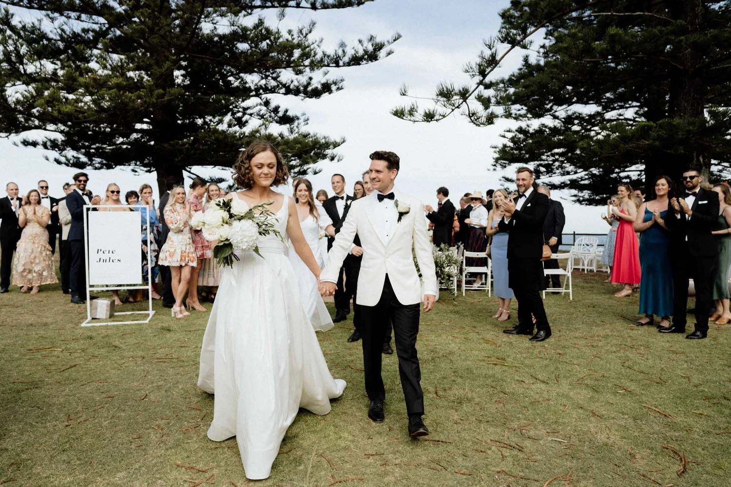 A bride and groom walking hand in hand at their outdoor wedding ceremony with guests clapping and smiling behind them.