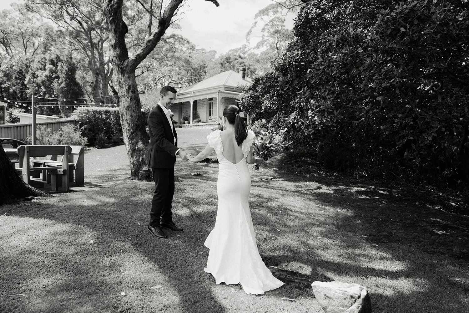 A black and white photo of a bride and groom holding hands outdoors, surrounded by trees and bushes in a garden setting during daytime.