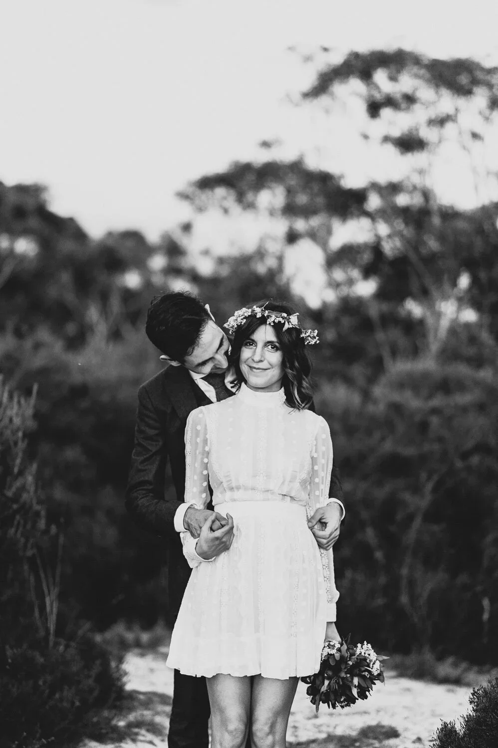 A black-and-white photo of a woman in a white dress with a floral crown holding a bouquet, standing outdoors, with a man in a suit standing behind her and holding her waist, in front of trees and a natural setting.