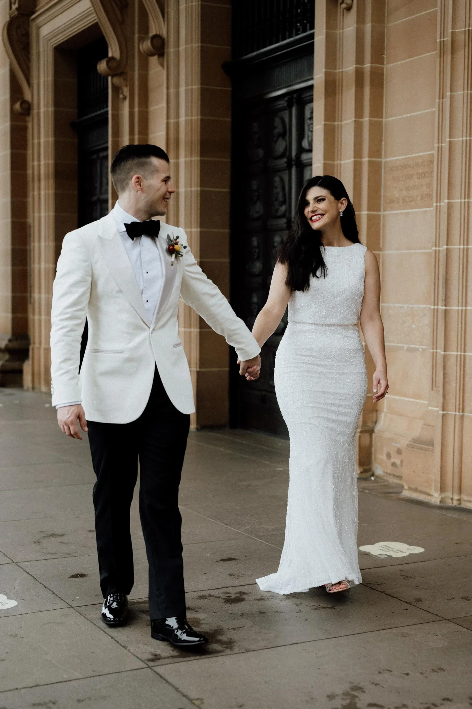 A newlywed couple holding hands and smiling at each other outside a building with ornate architectural details. The bride wears a white gown and the groom wears a white tuxedo jacket with black pants and a bow tie.