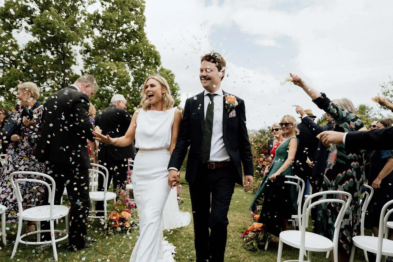 A newlywed couple walking hand-in-hand through a confetti-filled outdoor wedding ceremony. The bride is in a white dress with a joyful expression, and the groom is in a dark suit with a boutonnière, smiling. Guests are throwing confetti and celebrati