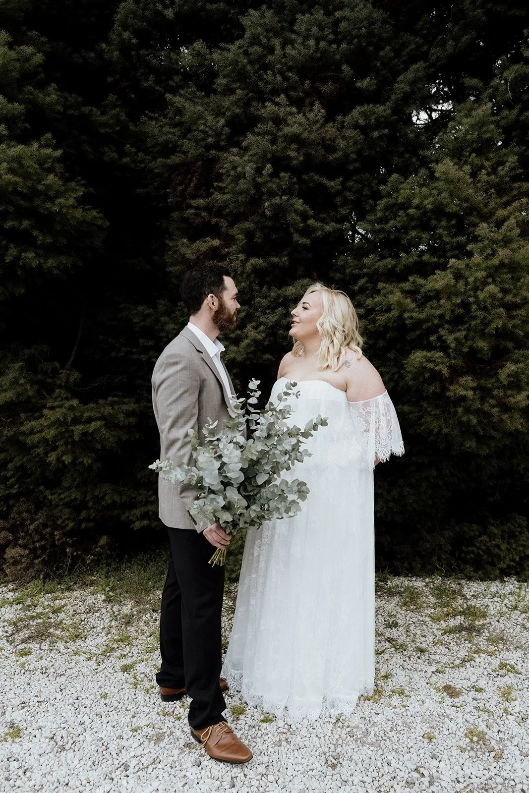 A bride and groom standing outdoors, gazing at each other, with large green trees in the background. The groom is holding a bouquet of eucalyptus.