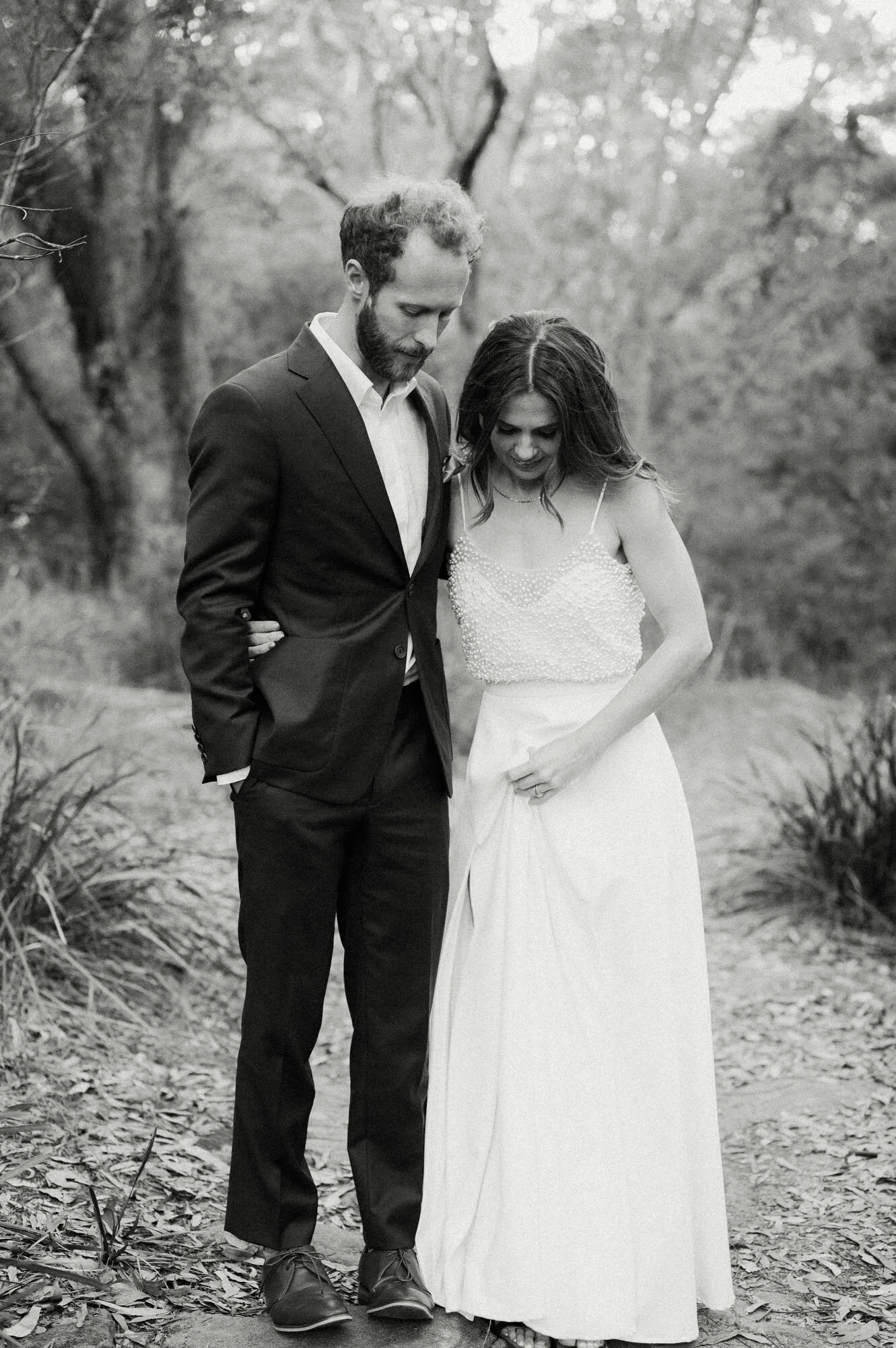 A black and white photograph of a couple outdoors, dressed in wedding attire; the man wears a suit and the woman wears a wedding dress with a beaded top. They are standing close together, looking down, with their heads bowed, in a natural setting wit