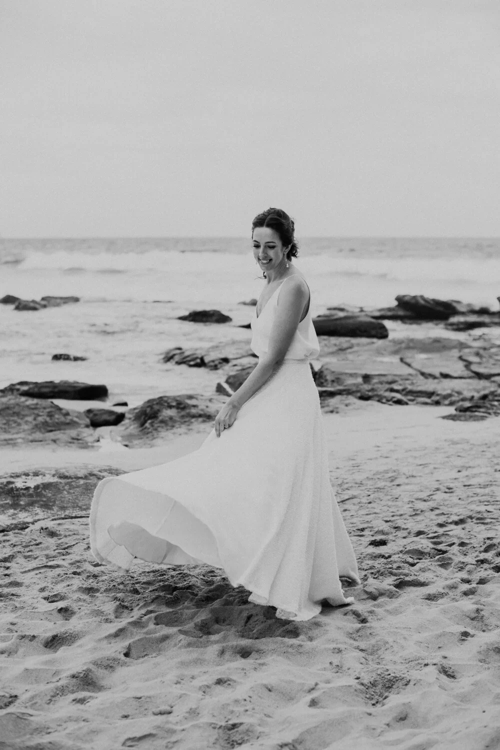 A woman in a long white dress standing barefoot on a sandy beach near rocks, smiling and looking down as she holds the side of her dress, with the ocean in the background.