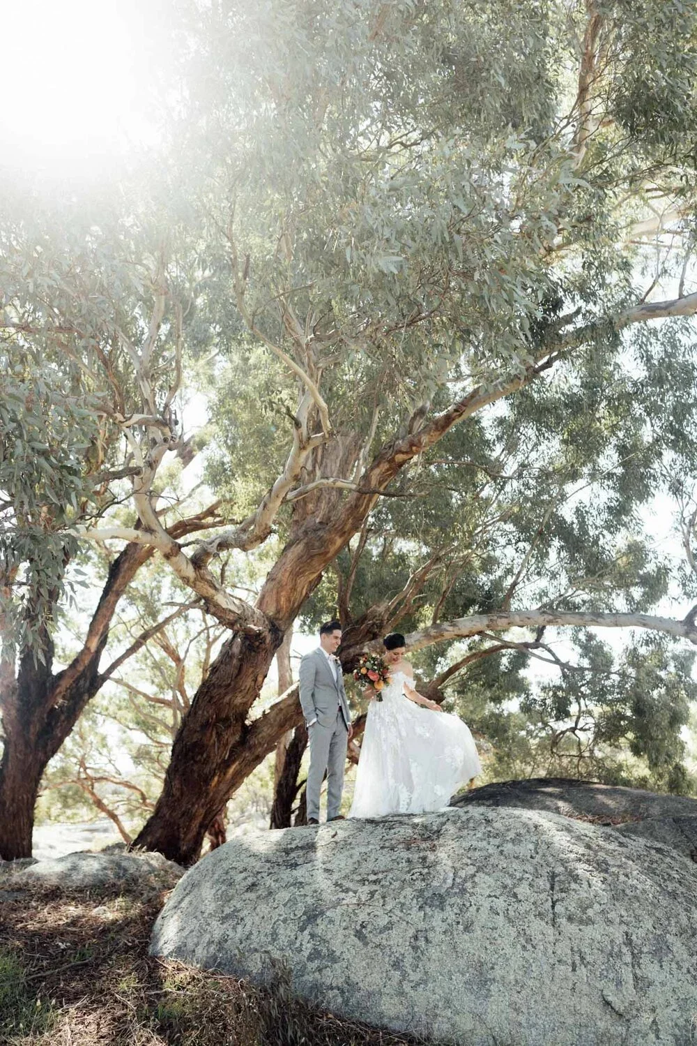 A bride and groom in wedding attire standing near a large tree and a rock outdoors, with sunlight filtering through the branches.