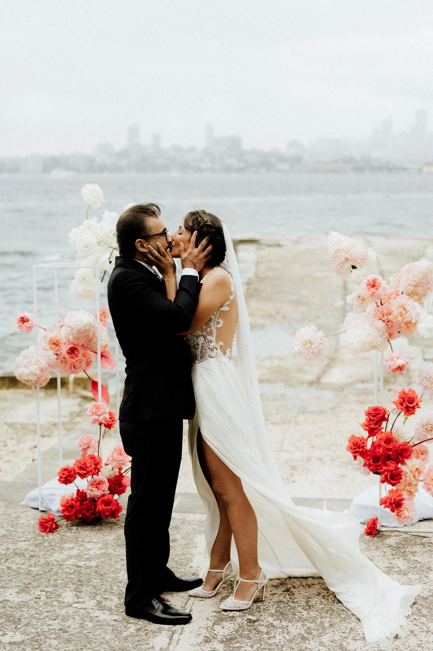 A couple dressed in wedding attire sharing a kiss on the beach surrounded by pink and white flowers with a city skyline and water in the background.