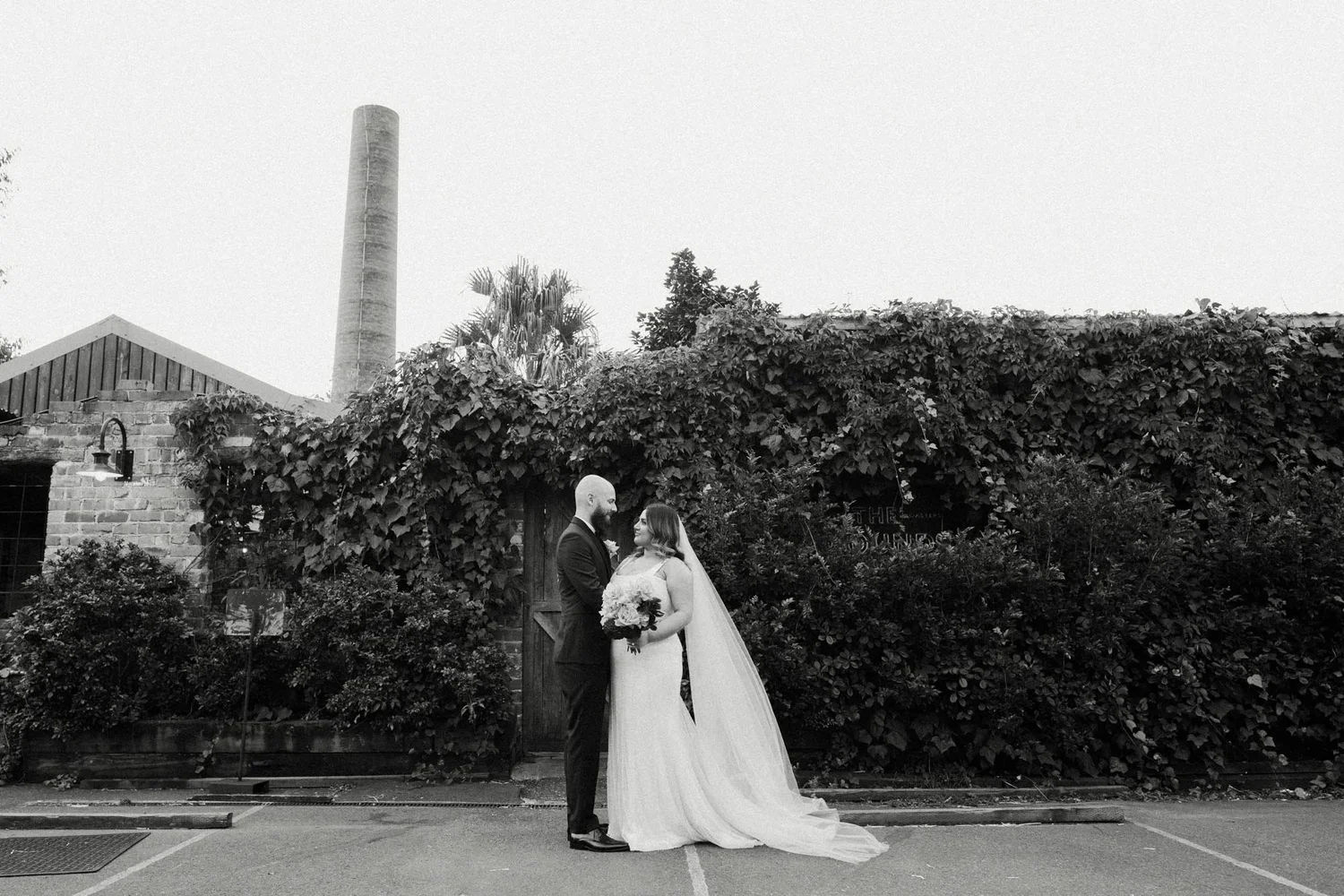Black and white photo of a bride and groom standing outdoors in front of a rustic wooden building with ivy and bushes. The groom is in a suit, and the bride is in a wedding dress holding a bouquet. They are looking at each other lovingly.