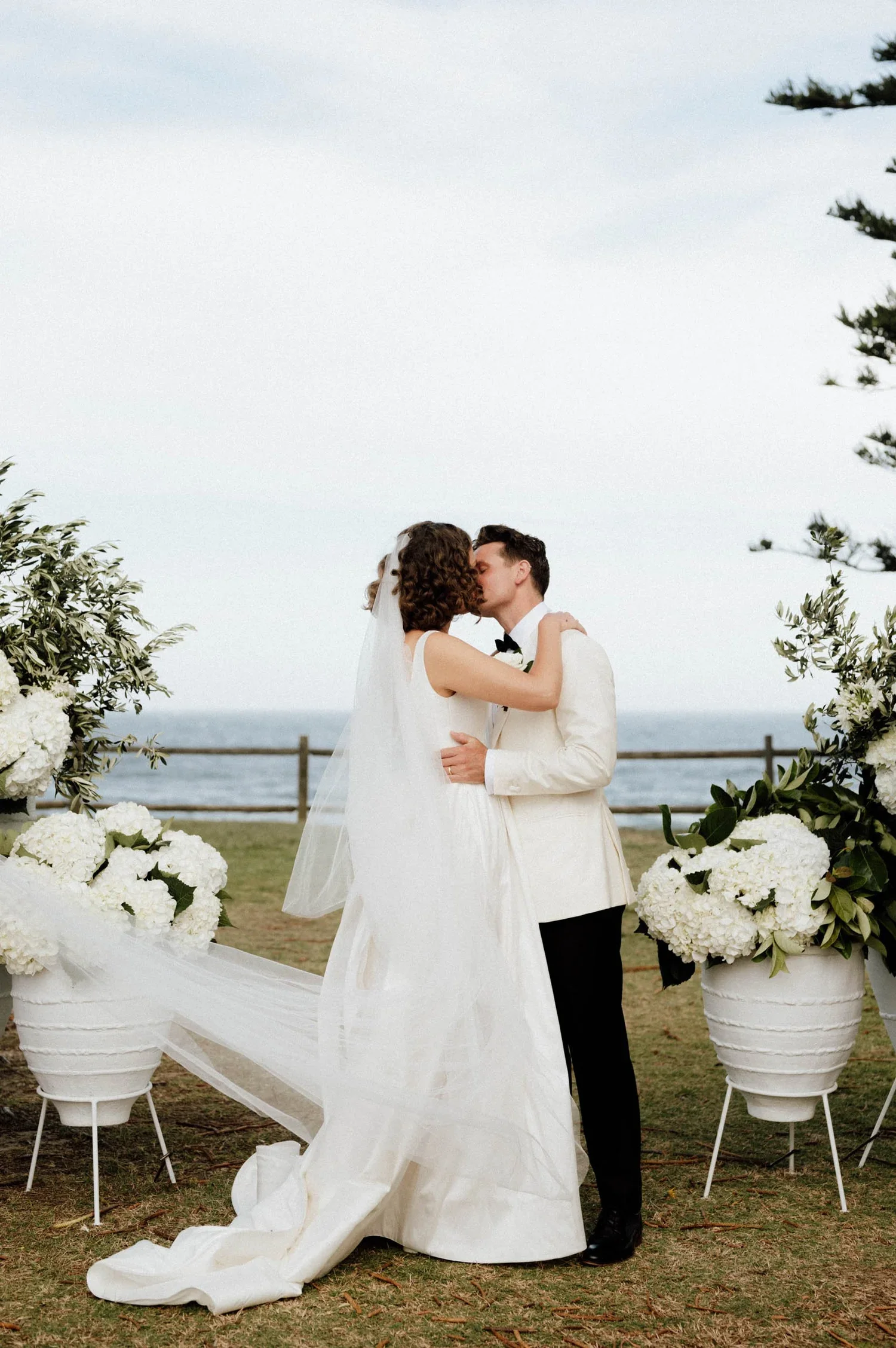 A bride and groom sharing a kiss at their outdoor wedding ceremony, with white flowers in large vases surrounding them and a wooden fence in the background.