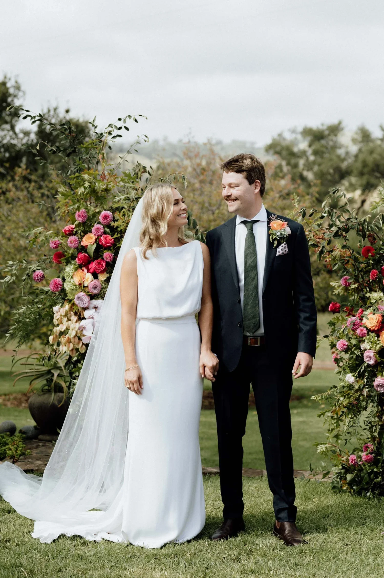 A bride and groom smiling and holding hands outdoors in front of floral arrangements. The bride is wearing a white wedding gown with a veil, and the groom is dressed in a black suit with a white shirt and a gray tie, with a boutonniere on his lapel.