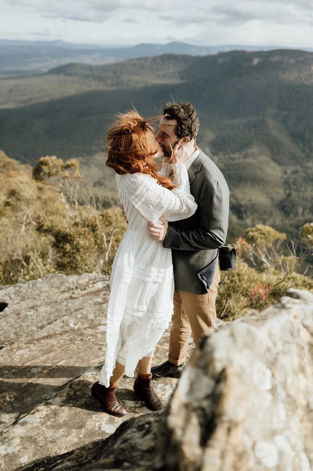 A couple embracing and kissing on a rocky mountain ledge, with a scenic view of rolling hills and mountains in the background.