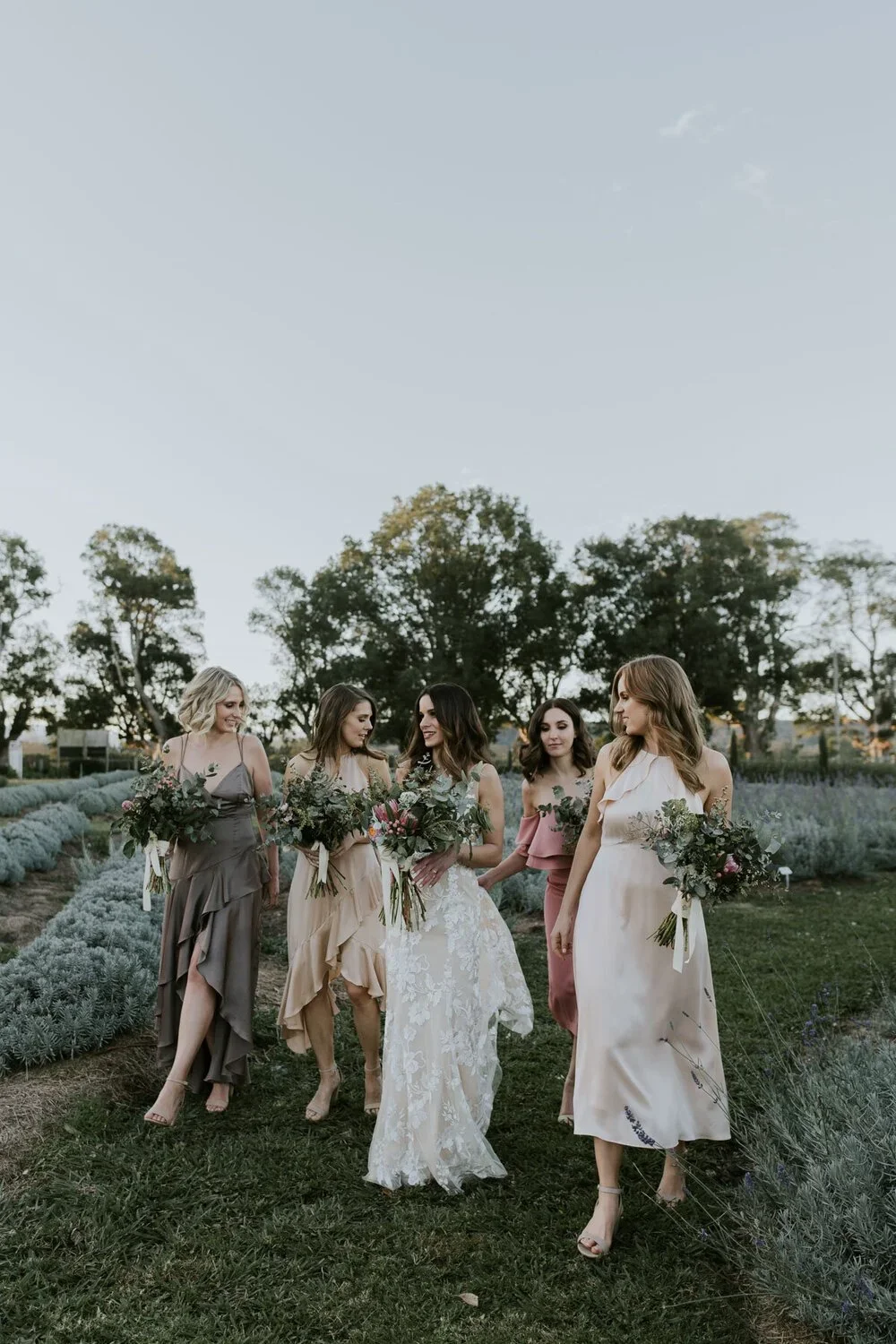 A bride walking with her bridesmaids in a garden during a wedding photoshoot, all holding bouquets of flowers.