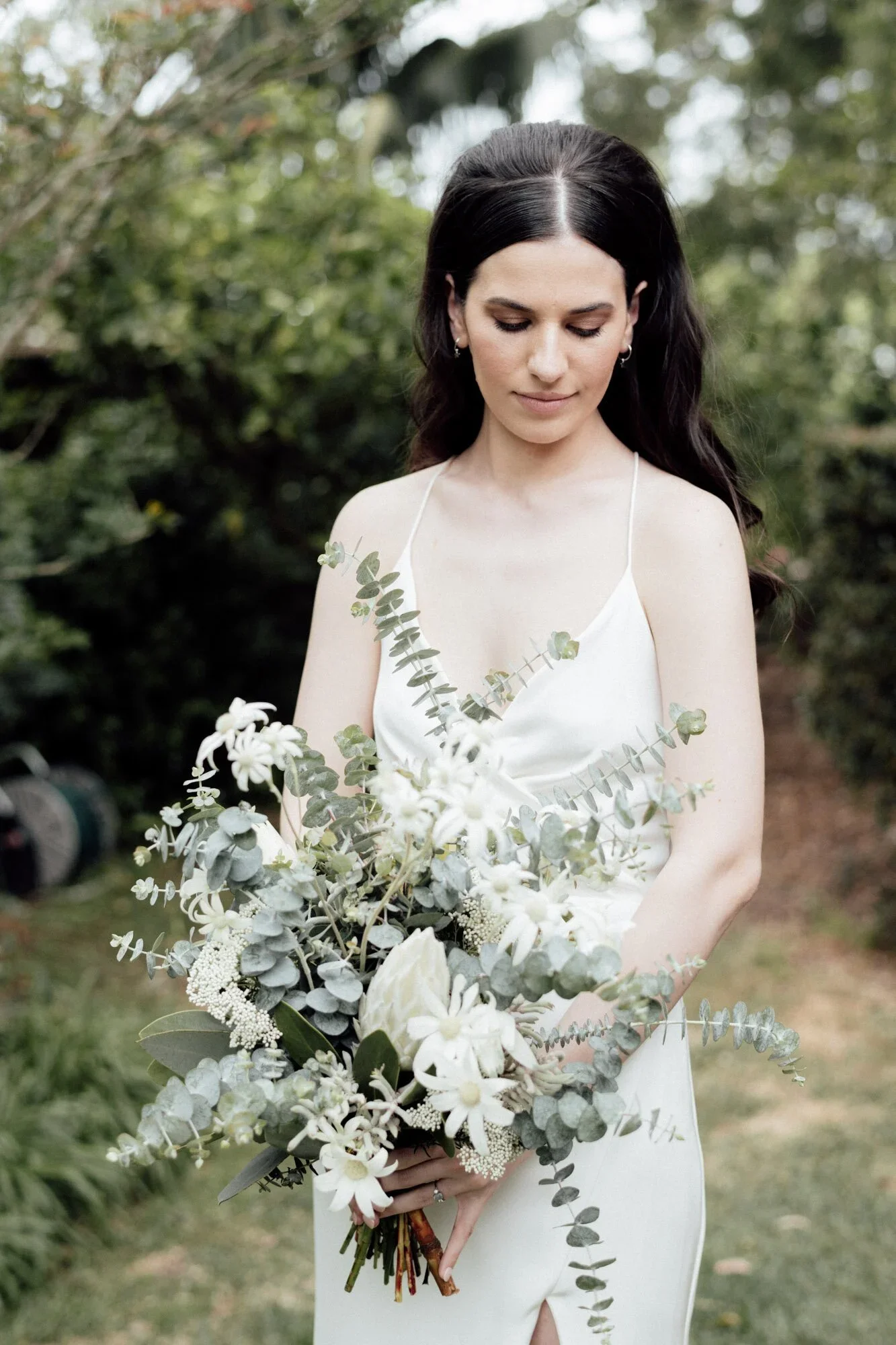 A woman in a white dress holding a large bouquet of white and green flowers, standing outdoors with trees in the background.