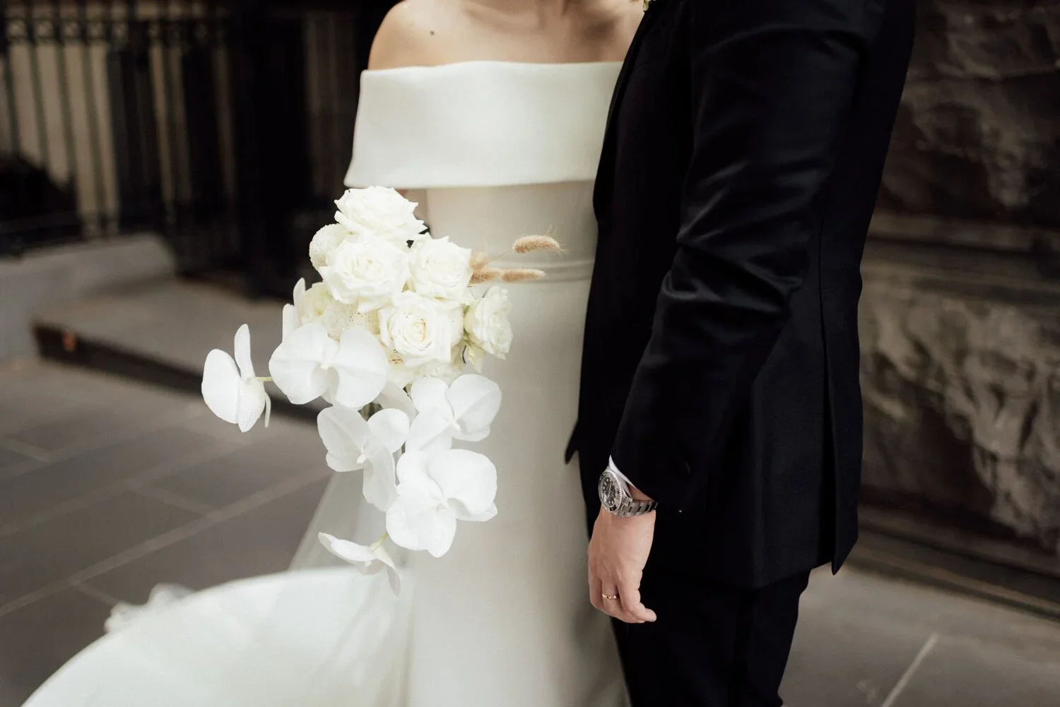 A bride and groom standing close together. The bride wears an off-the-shoulder white wedding gown and holds a bouquet of white roses and orchids. The groom is dressed in a black suit with a silver watch.