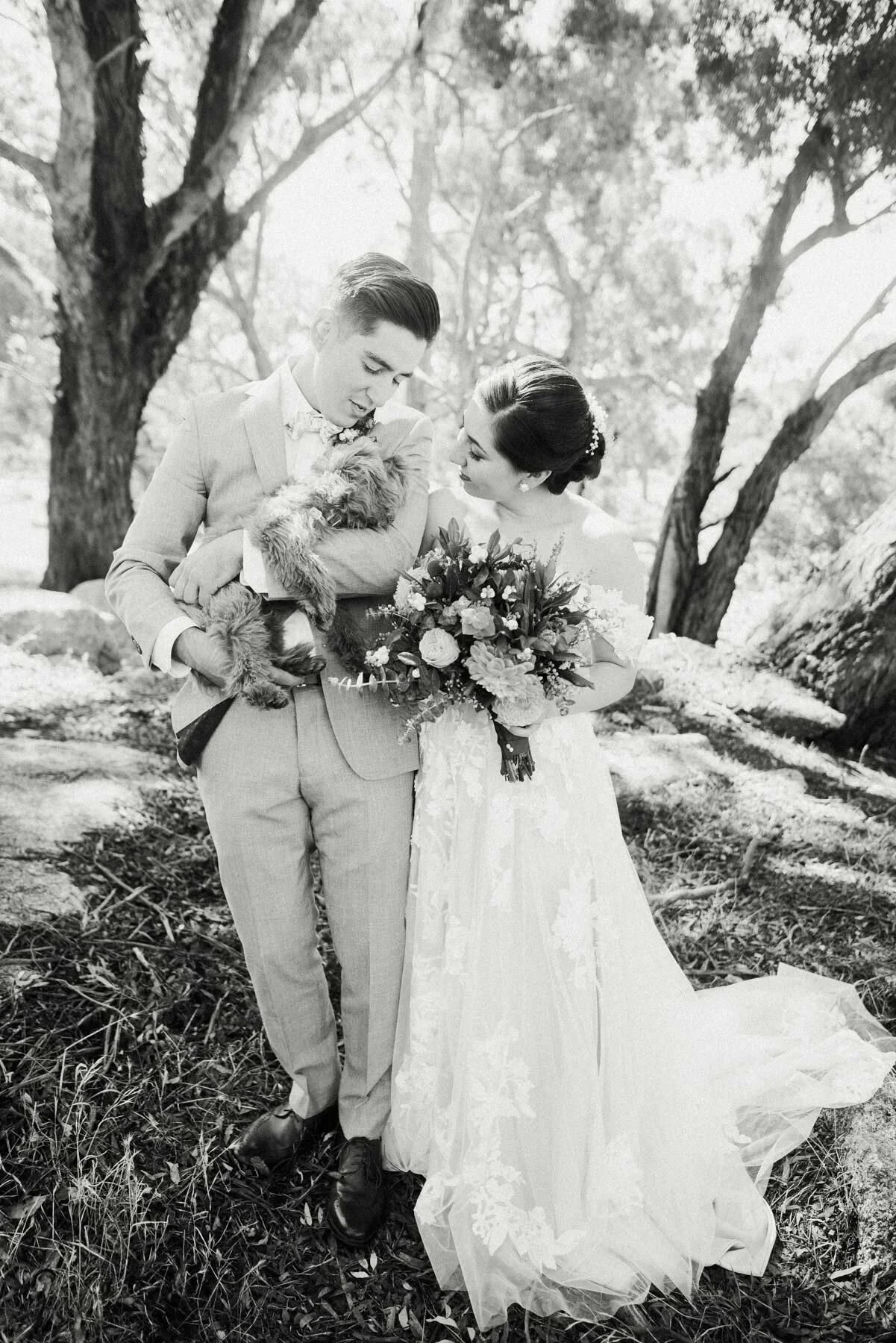 A black-and-white photograph of a bride and groom standing outdoors among trees. The groom, in a light suit with a bow tie, holds a small dog. The bride, in a lace wedding gown holding a bouquet of flowers, looks at the groom. The setting appears nat