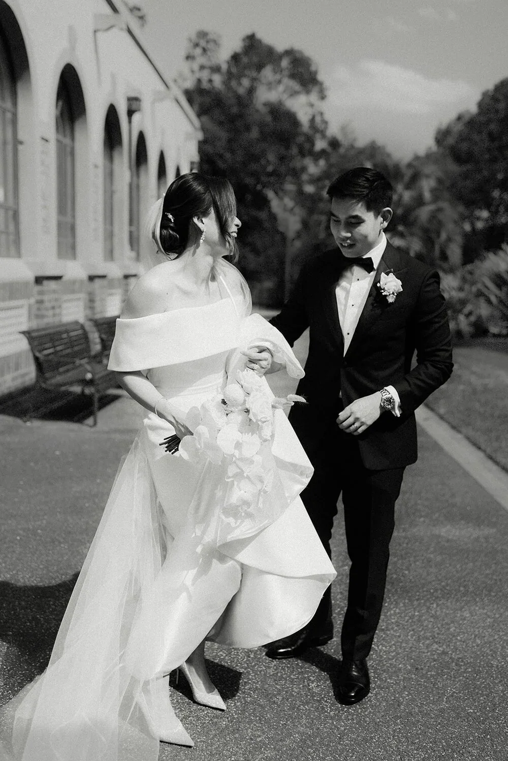 A bride and groom share a moment outdoors, the bride in a white wedding gown holding a floral bouquet and the groom in a tuxedo with a boutonniere, smiling at each other during their wedding.