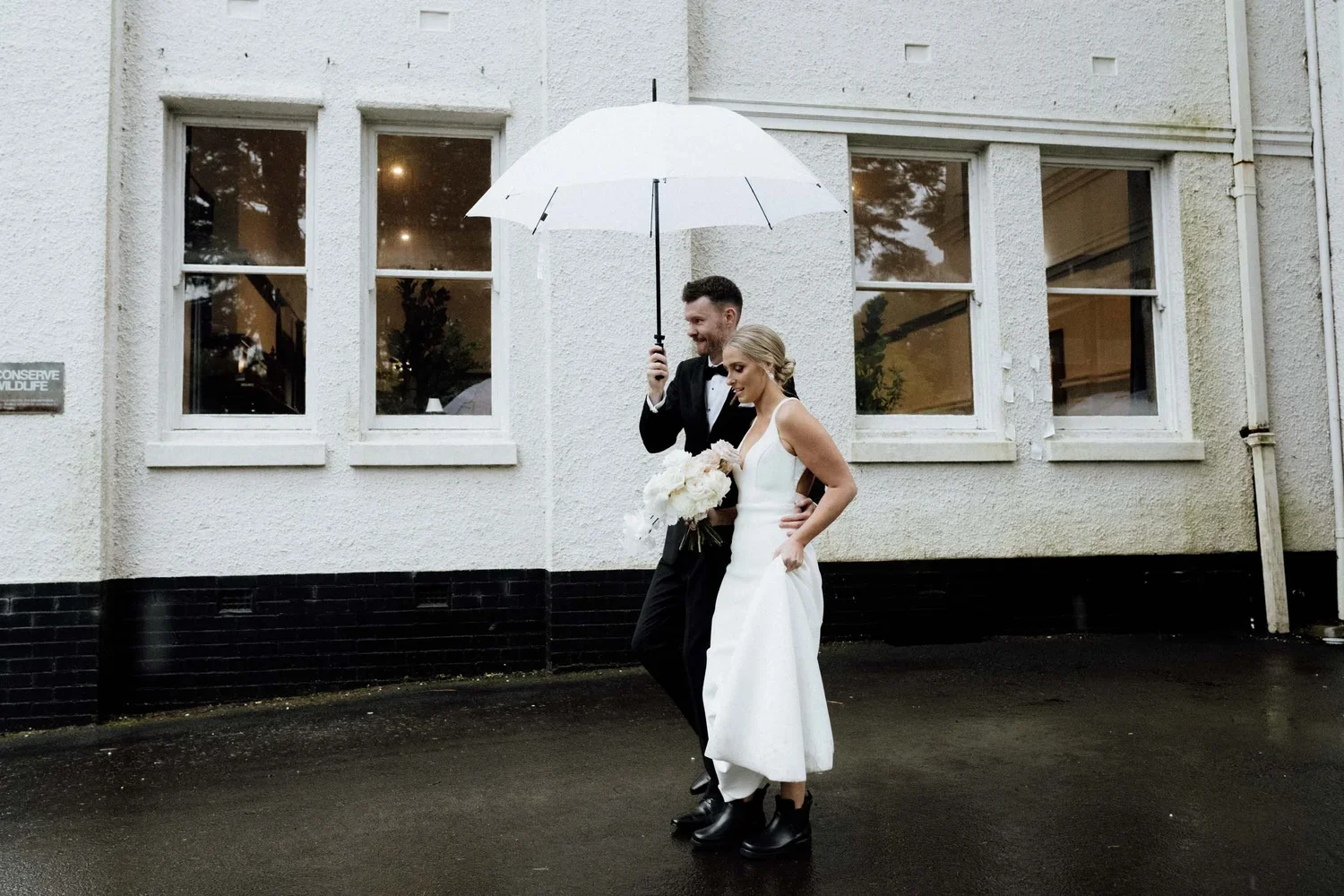 A bride and groom stand under a white umbrella outside a building, with the groom holding the umbrella and the bride holding a bouquet of flowers, as they enjoy a rainy wedding day.